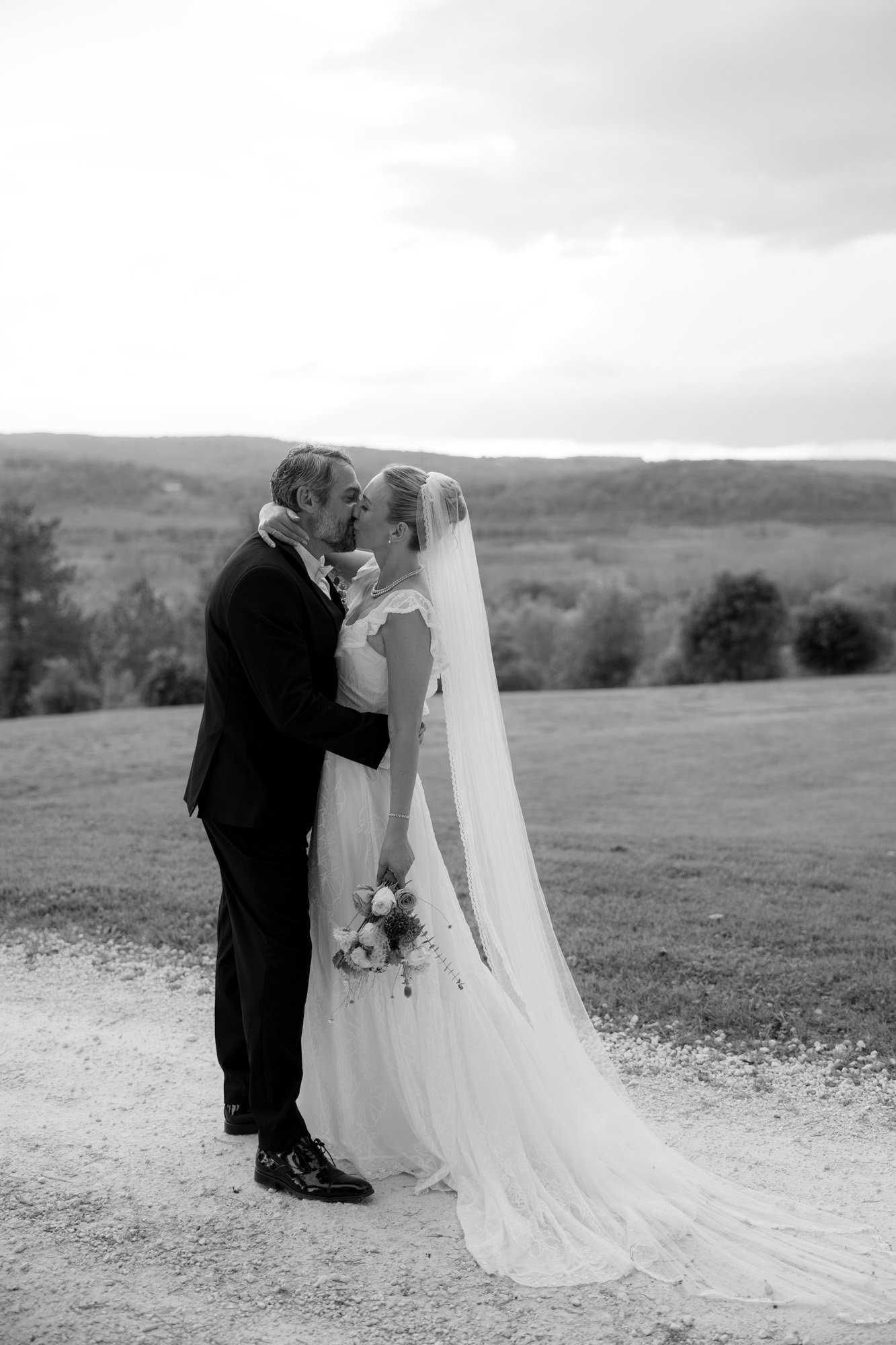 A bride and groom stand close together outdoors, dressed in formal wedding attire, with the bride holding a bouquet and a landscape in the background.