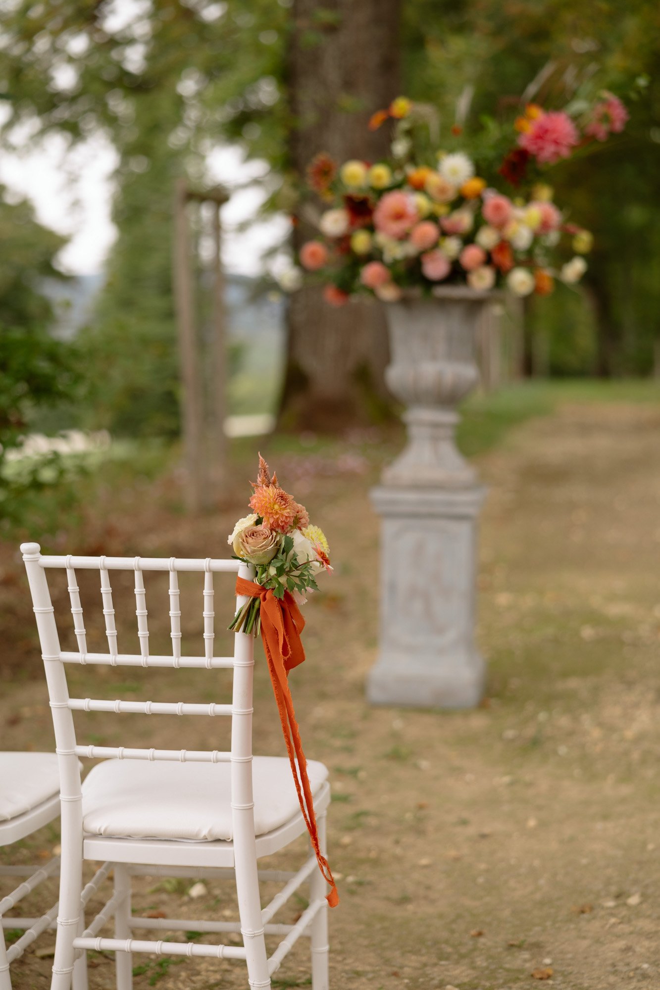 A white chair decorated with flowers and an orange ribbon stands outdoors near a large floral arrangement on a pedestal. Autumn France wedding.