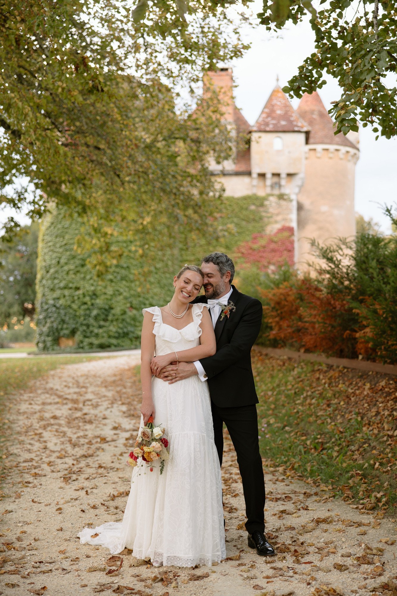 A bride in a white dress and a groom in a black suit stand on a leaf-covered path, embracing and smiling, with a castle and greenery in the background. Autumn France wedding.