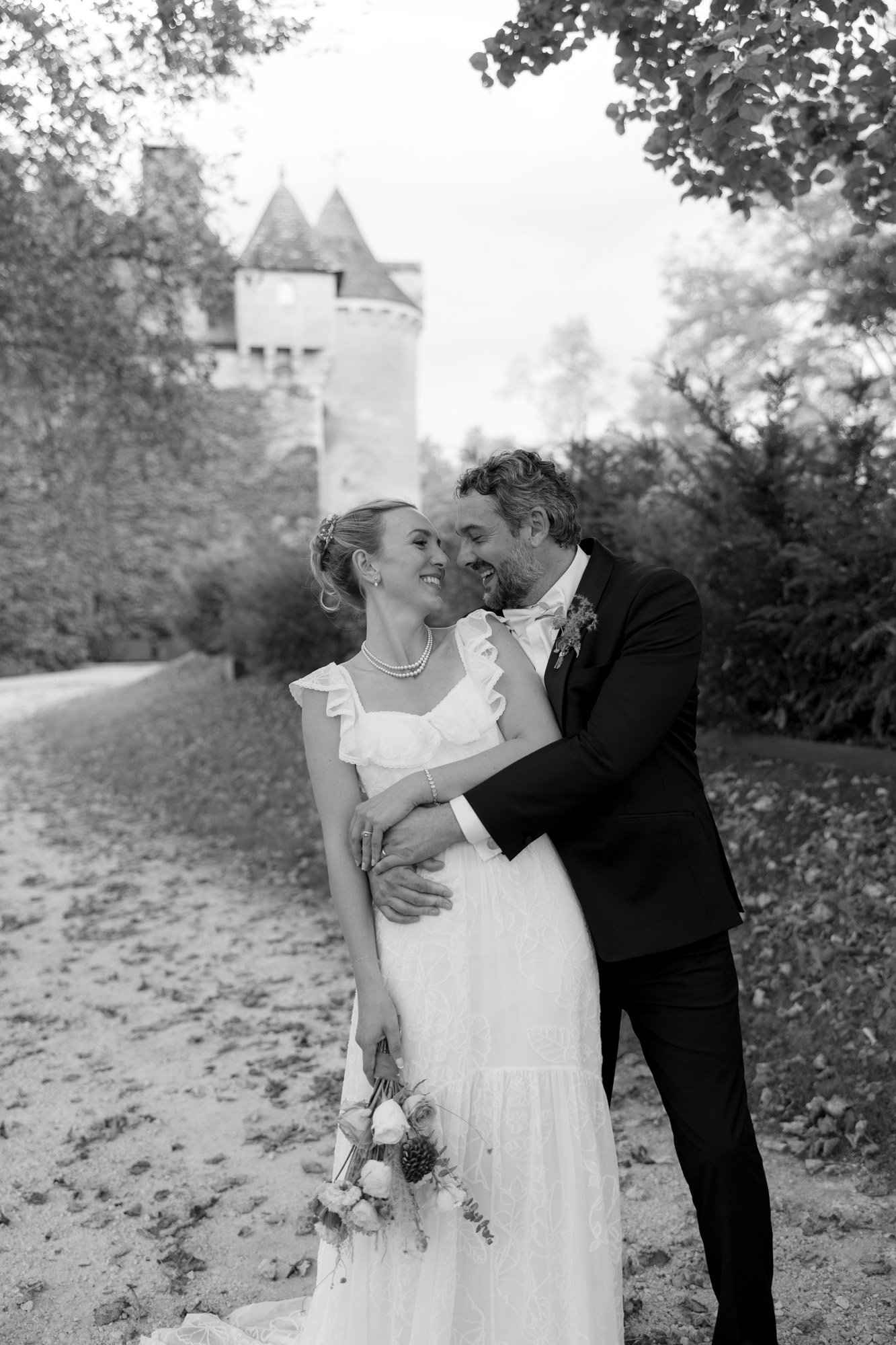 A bride and groom stand on a tree-lined path, smiling and embracing, with a bouquet in the brides hand and a castle in the background.