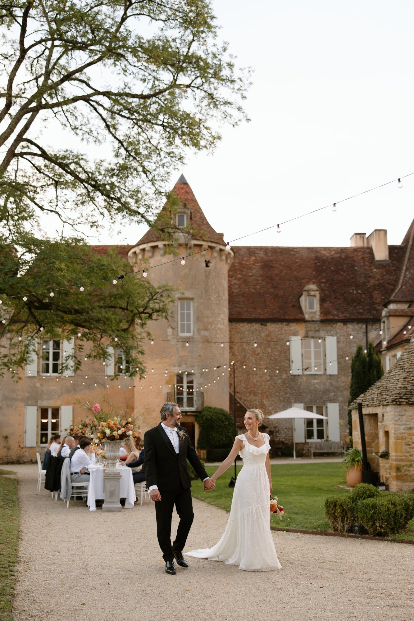 A bride and groom walk hand in hand outside a historic stone building, with wedding guests seated at outdoor tables and string lights overhead. Autumn France wedding.
