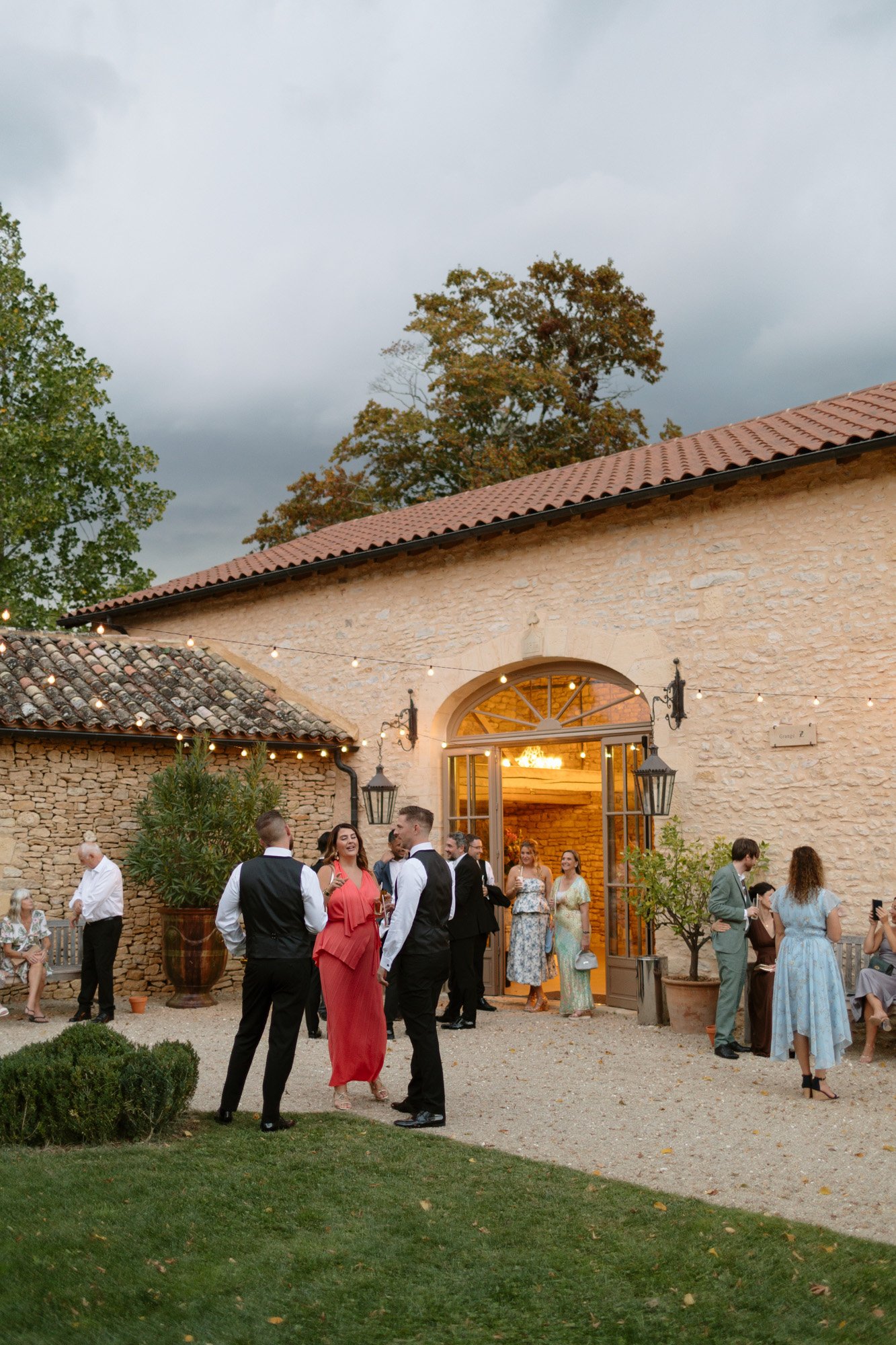 Guests in formal attire mingle outside a rustic stone building with string lights overhead, on a cloudy day. Autumn France wedding.