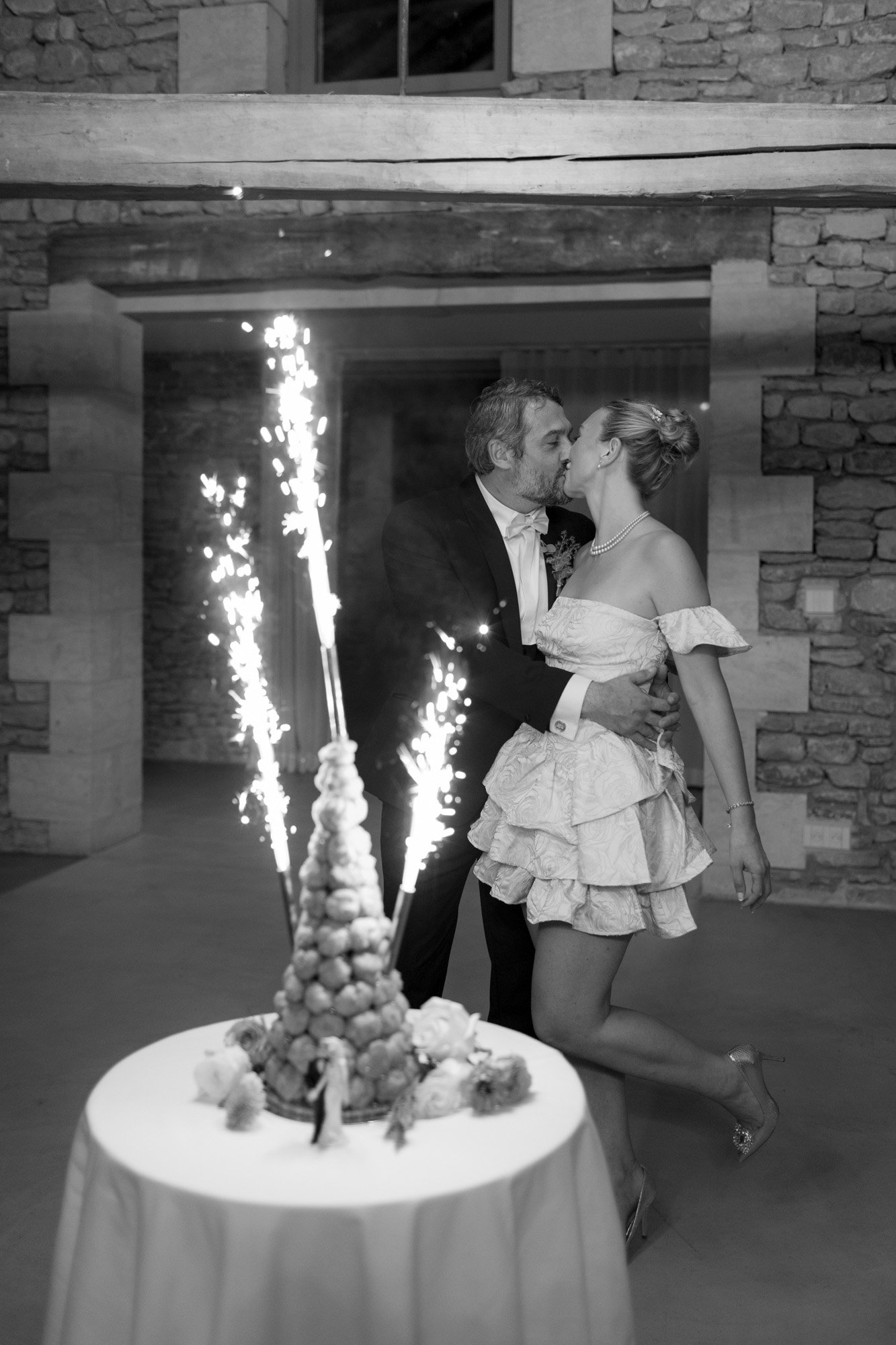 A couple dressed in formal attire kisses near a croquembouche dessert with sparklers, in a stone-walled room.