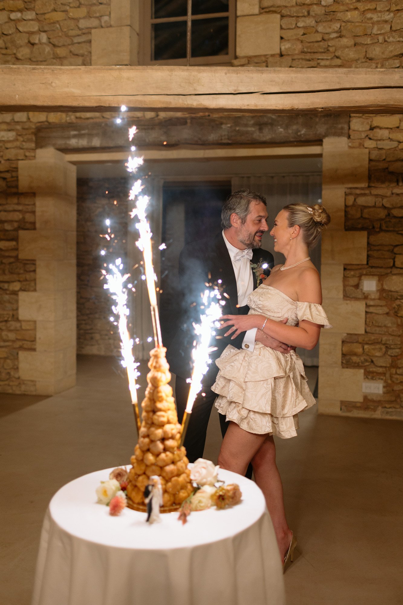 A couple dressed formally stands close together, smiling near a croquembouche dessert topped with sparklers on a table in a rustic stone-walled room. Autumn France wedding.
