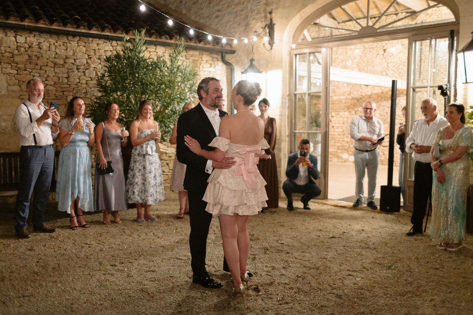 A couple dances in the center of a rustic indoor space while a group of people dressed formally watch and take photos.