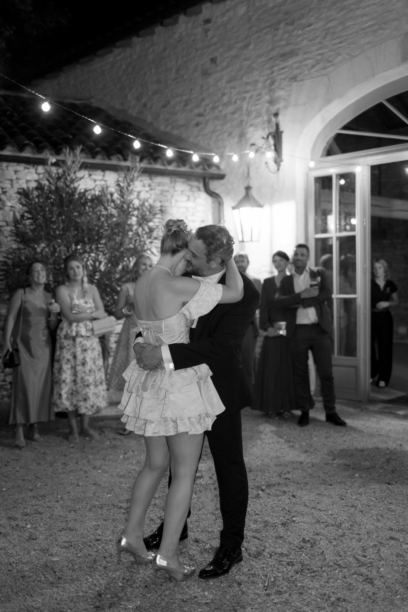 A couple embraces and dances closely outdoors at night, surrounded by people watching and clapping at a formal event.