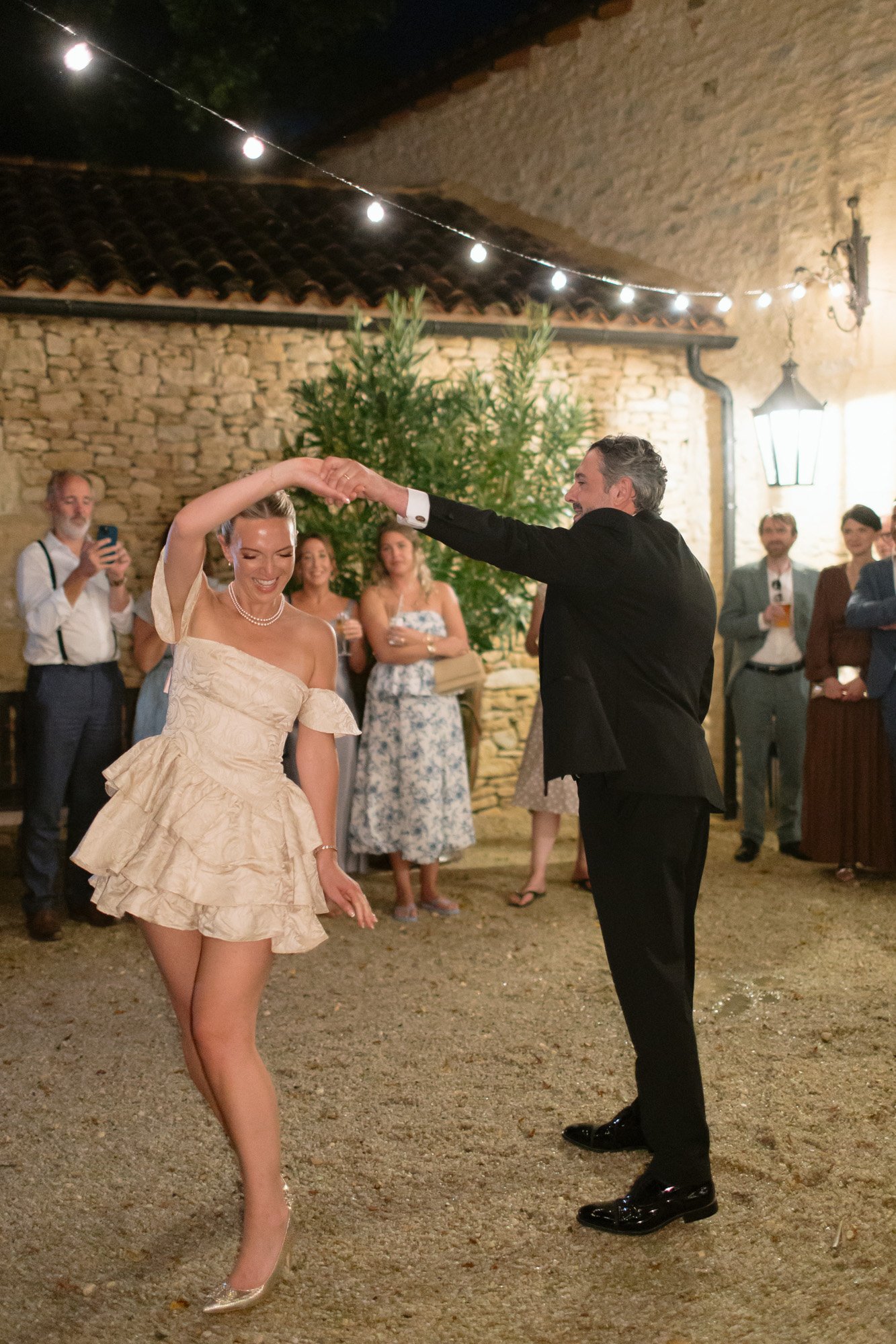 A man in a suit twirls a smiling woman in a short, off-the-shoulder dress while guests watch at an outdoor evening event. Autumn France wedding.