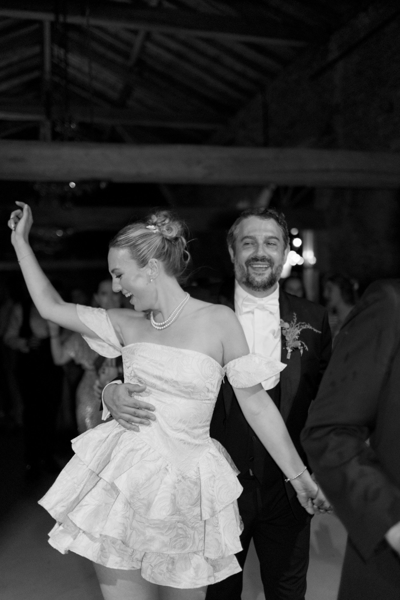 A woman in a short, off-shoulder dress dances with a man in a suit and bow tie at an indoor event. Both are smiling. The setting appears to be a dimly lit venue with exposed beams.