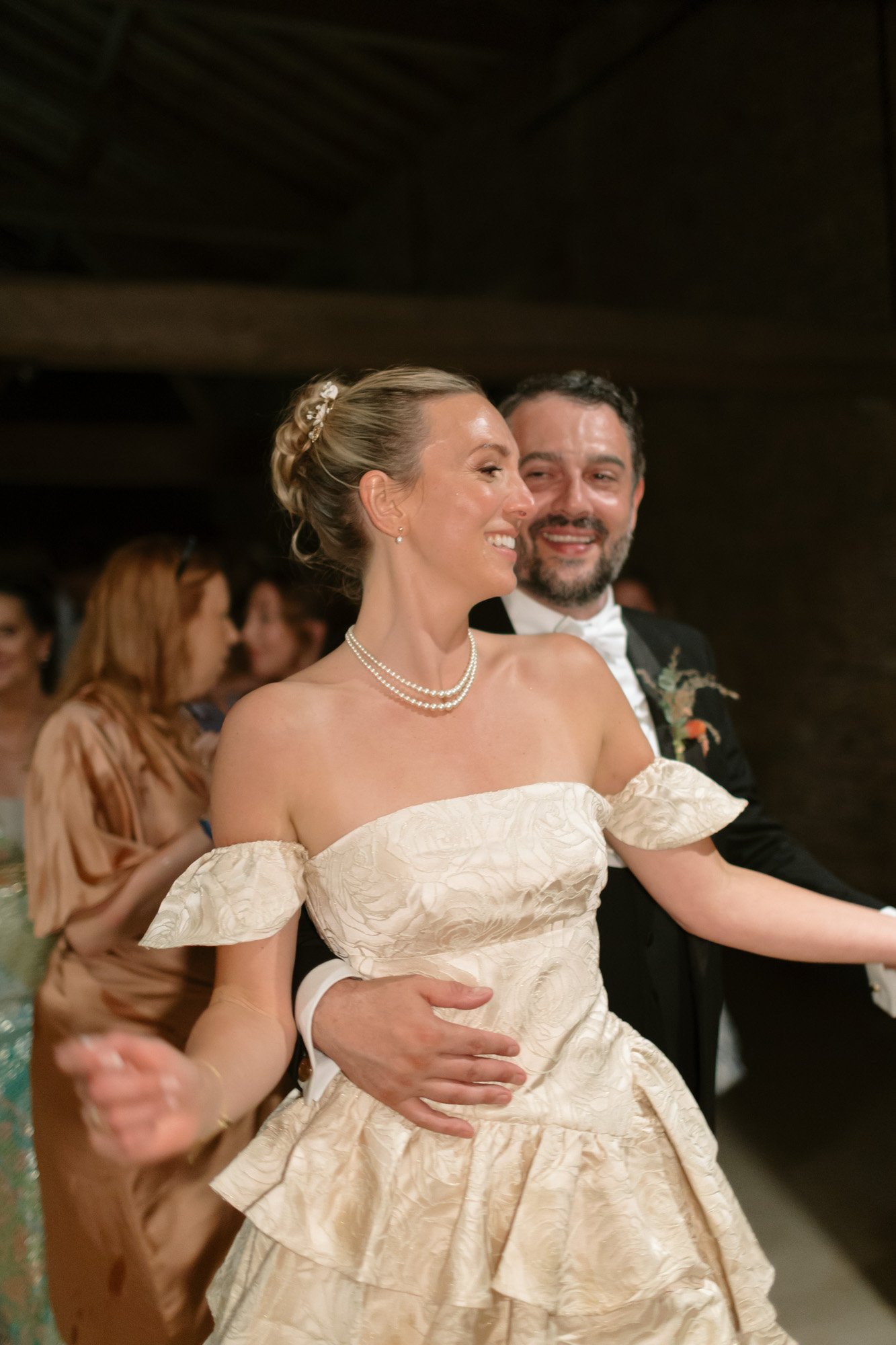 A bride in an off-shoulder wedding dress and a groom in a suit smile and dance together at a wedding reception, surrounded by guests.