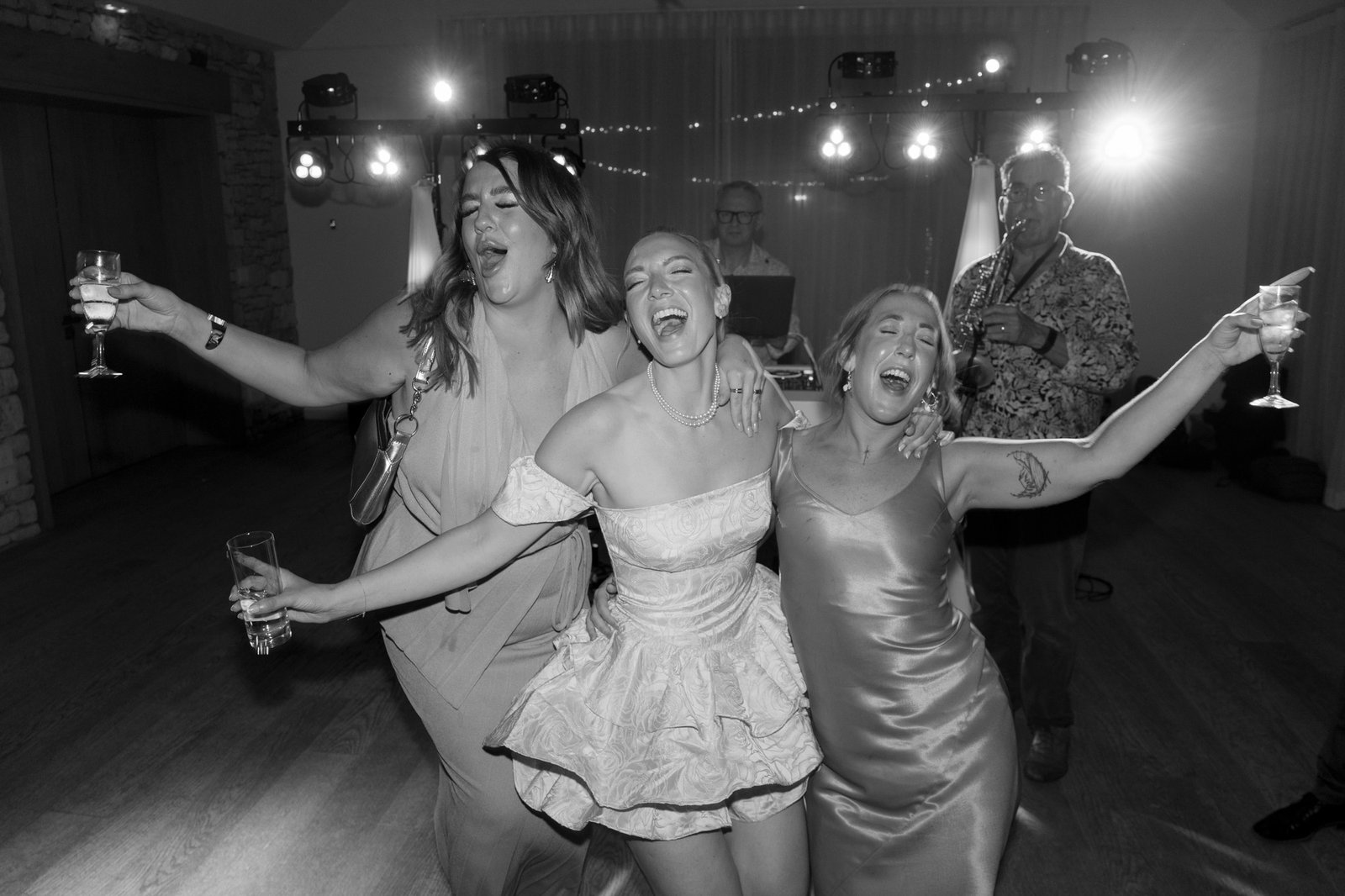 Three women in dresses sing and dance together at a party, holding drinks, while two men stand in the background near a DJ setup. The scene is in black and white.