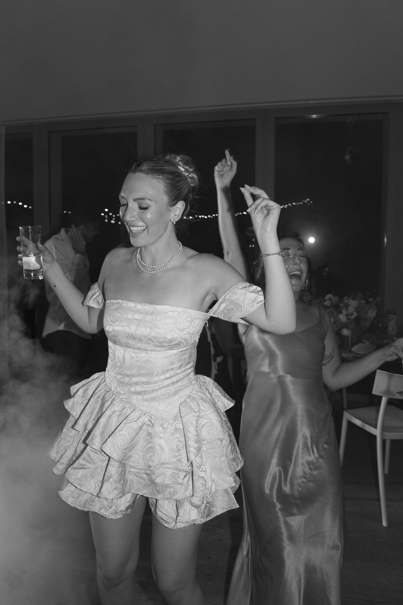 Two women dance and smile at an indoor event, one holding a drink, with visible smoke and string lights in the background.