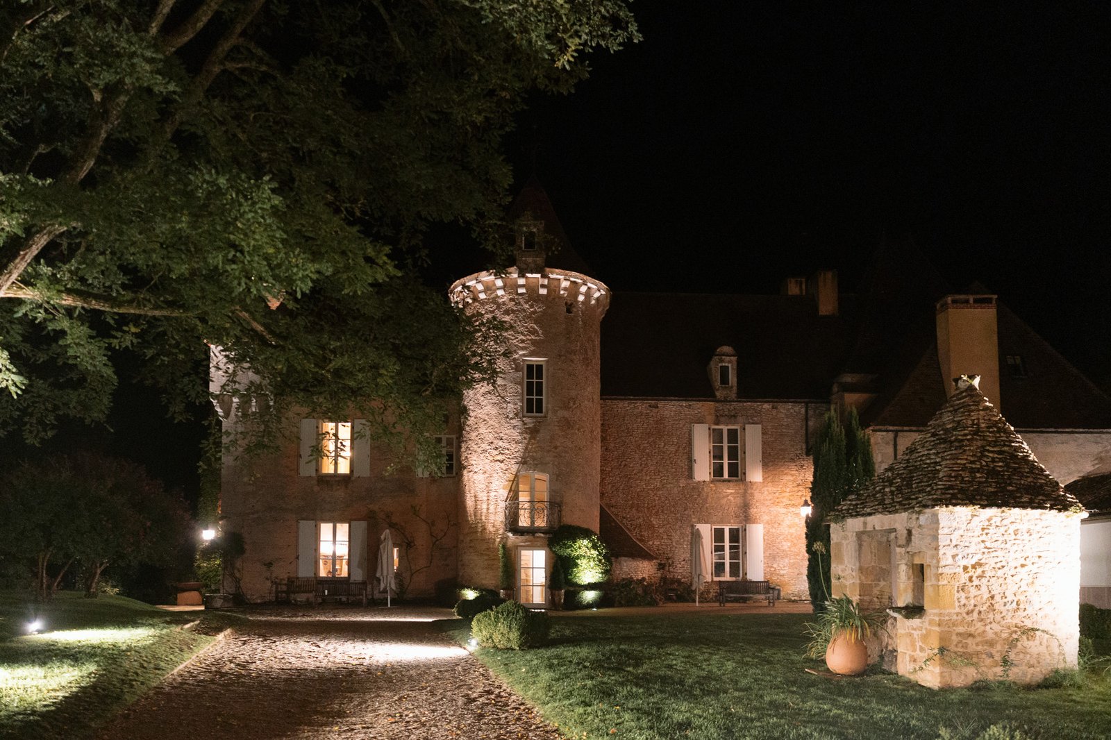 A stone chateau with round tower and warm lighting stands at night, surrounded by trees and landscaped grounds.