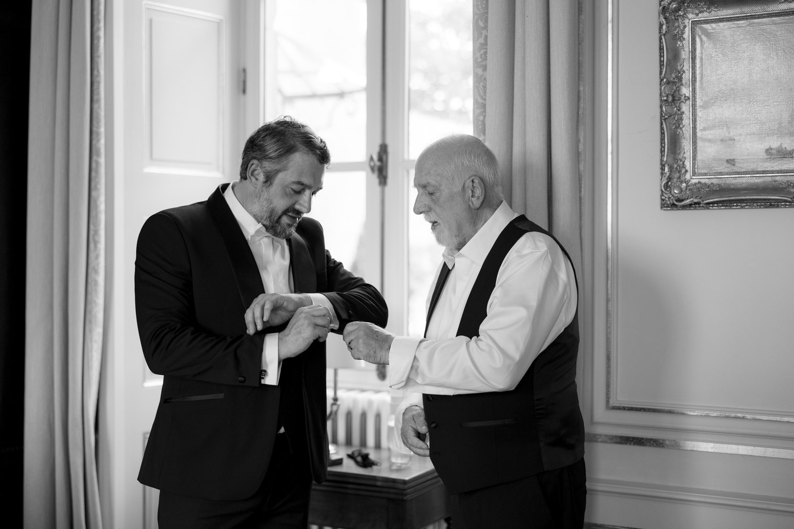 Two men in formal attire stand indoors, adjusting their cuffs while facing each other. One appears older with white hair, the other younger with dark hair. The setting is elegant and well-lit.
