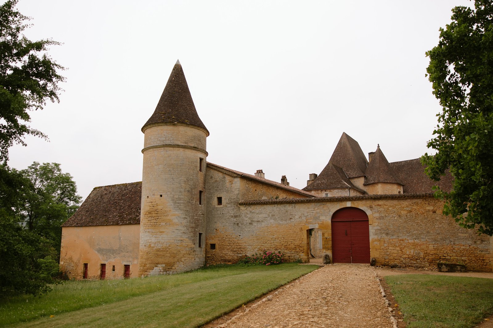 Stone chateau with conical towers, sloped roofs, and a red arched wooden gate, surrounded by greenery and a gravel path.