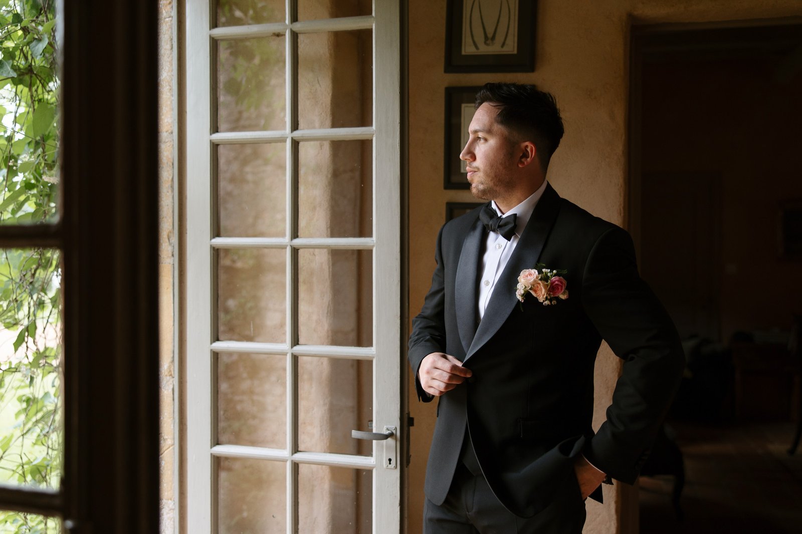 A man in a black tuxedo with a boutonniere stands indoors by a glass door, looking outside with one hand in his pocket.