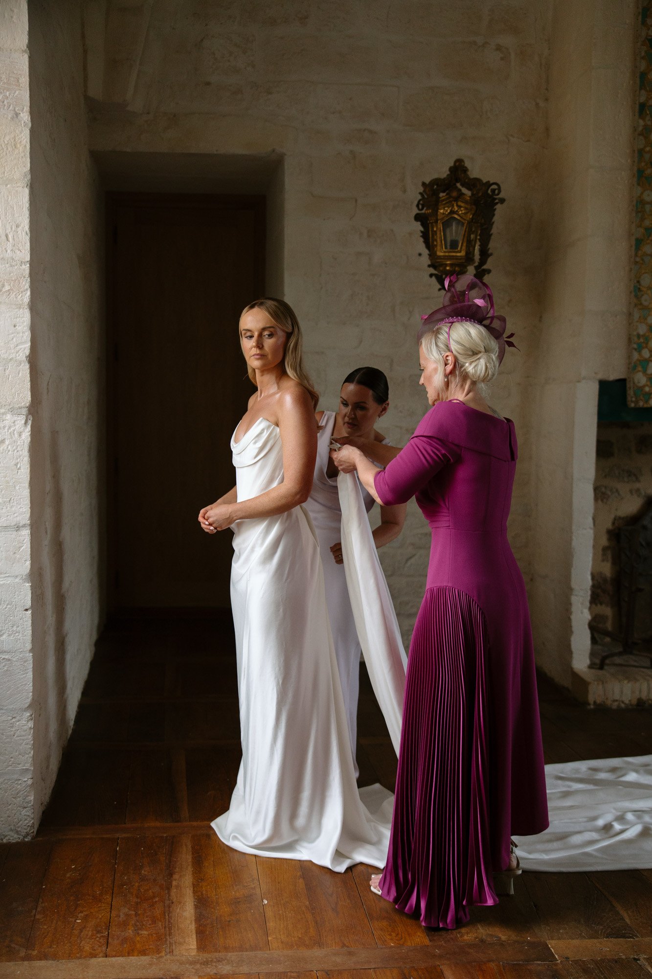 A bride in a white gown stands as two women, one in a purple dress and hat, adjust her dress in a room with stone walls and wooden floors.