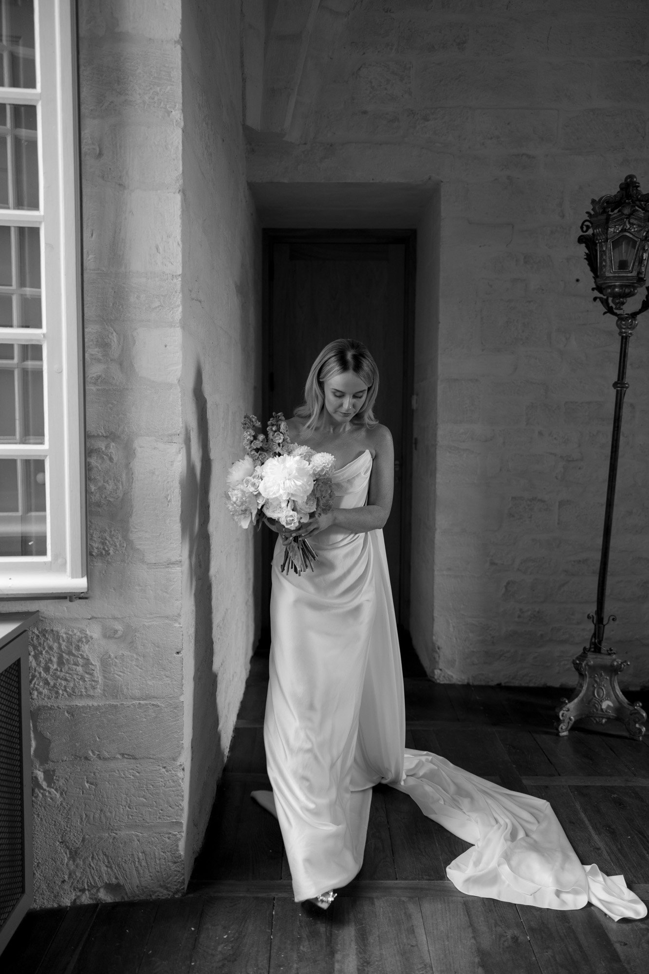 A woman in a long, flowing gown holds a bouquet of flowers and walks indoors along a stone wall, with a window and ornate lamp visible. Chateau de la Bourlie wedding.