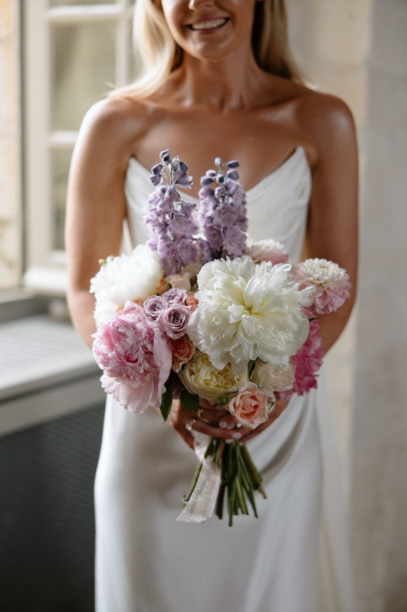 A woman in a white dress holds a bouquet of pastel-colored flowers, including white peonies, pink roses, and purple delphiniums. Chateau de la Bourlie wedding.