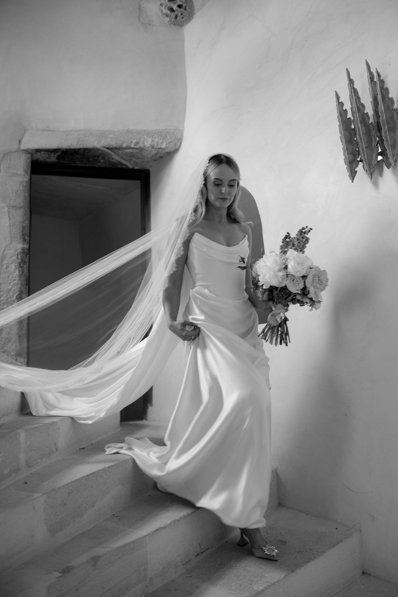 A bride in a long, flowing gown and veil descends a stone staircase while holding a bouquet of flowers.