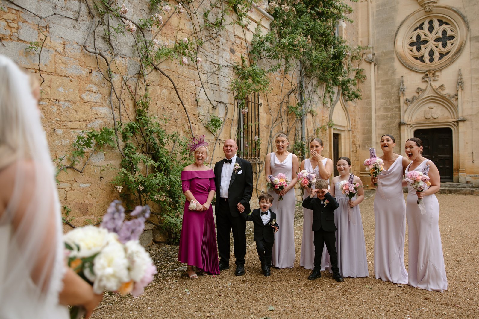 A wedding party stands outside a stone building, smiling at the bride, who holds a bouquet in the foreground. Chateau de la Bourlie wedding.