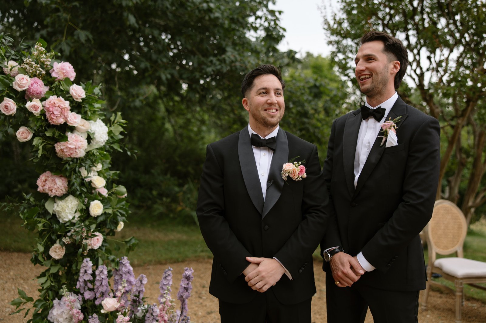 Two men in black tuxedos stand by a floral arrangement outdoors, smiling, with greenery and a chair in the background. Chateau de la Bourlie wedding.