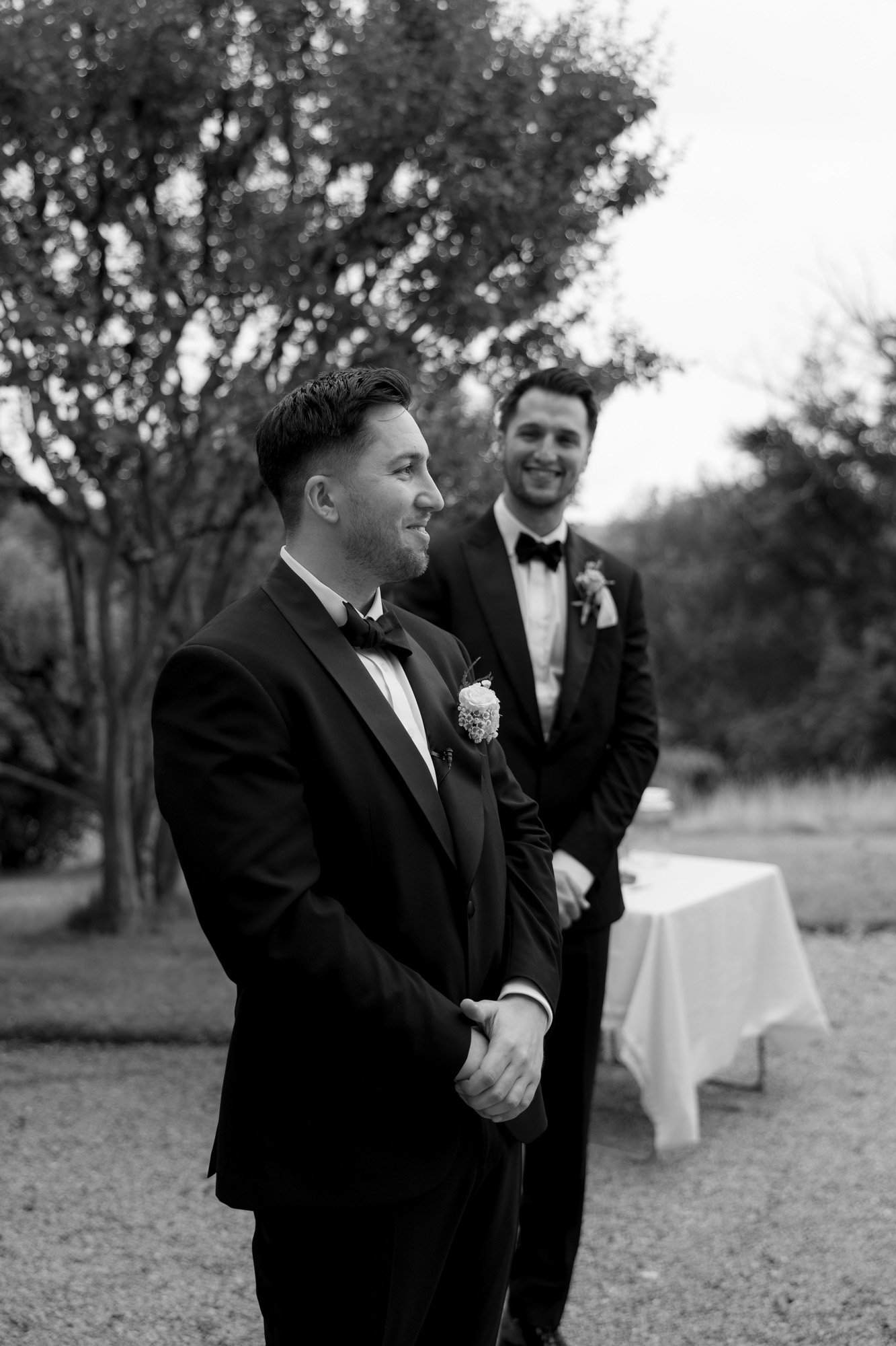 Two men in tuxedos stand outdoors in front of a table and trees, with one in the foreground and one smiling in the background. The setting appears formal and is in black and white.