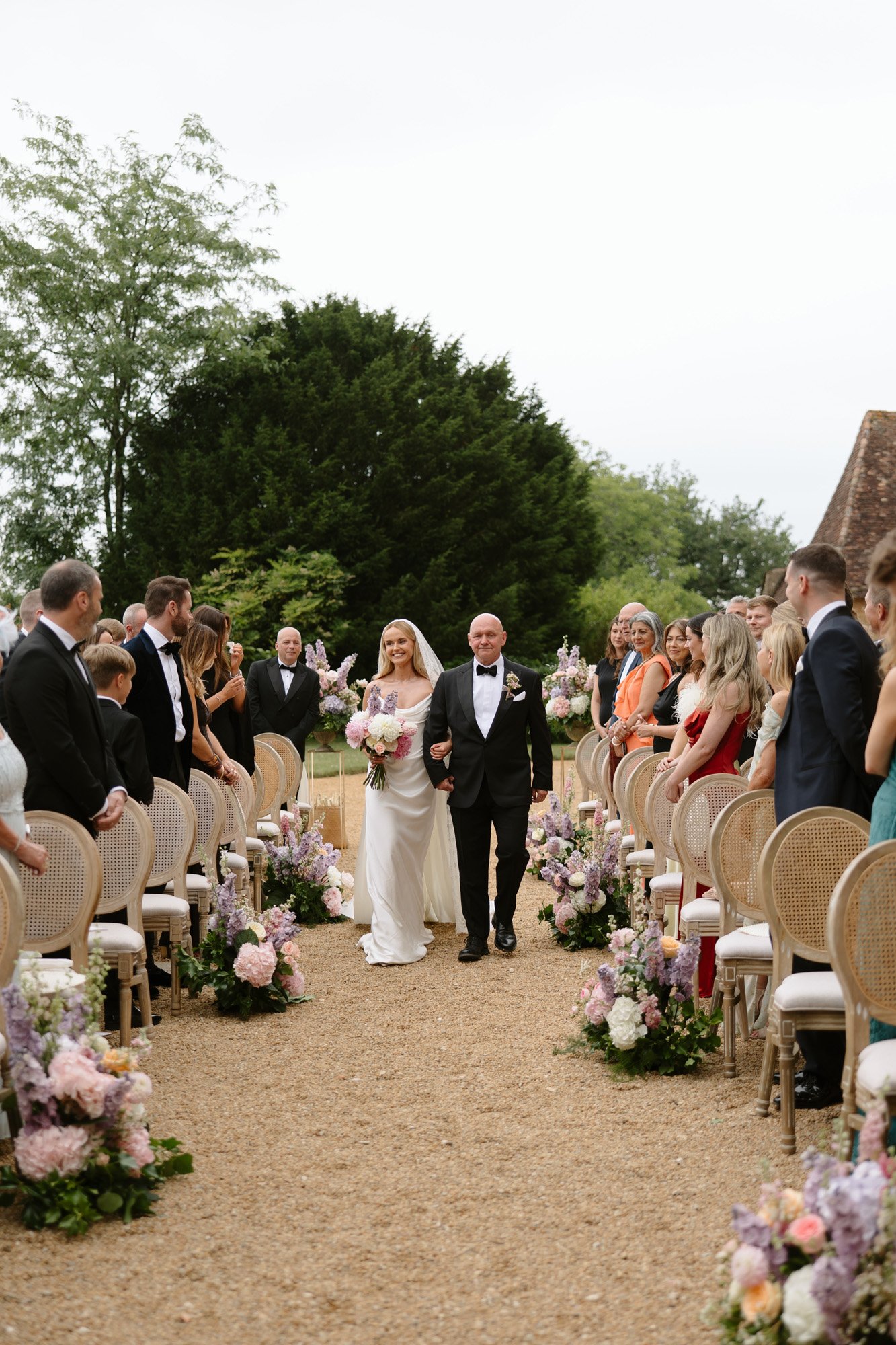 A bride in a white dress walks down an outdoor aisle with a man in a tuxedo, surrounded by seated guests and floral arrangements. Chateau de la Bourlie wedding.