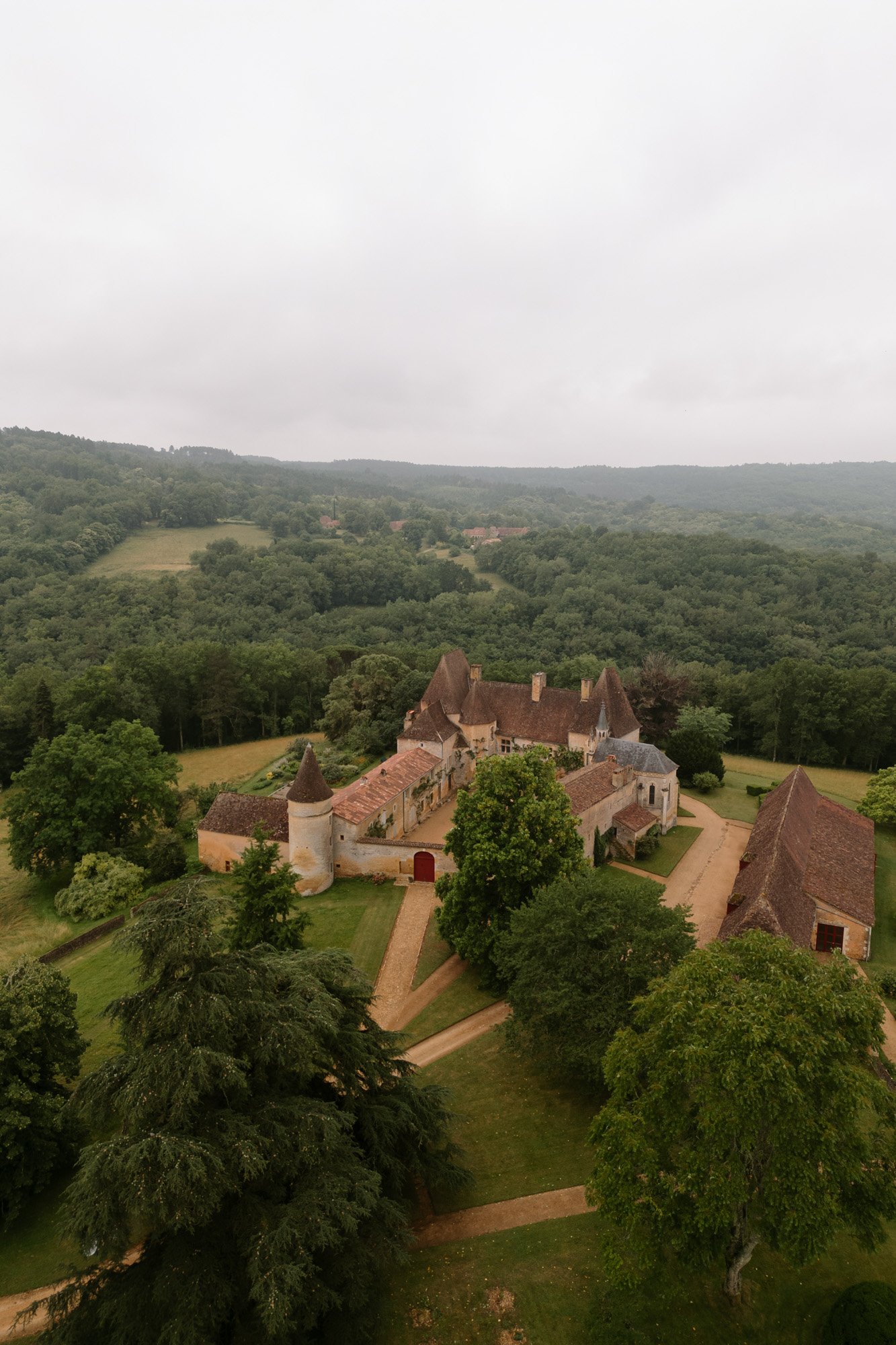 Aerial view of a historic stone estate with red roofs and towers, surrounded by trees and rolling green hills under a cloudy sky. Chateau de la Bourlie wedding.