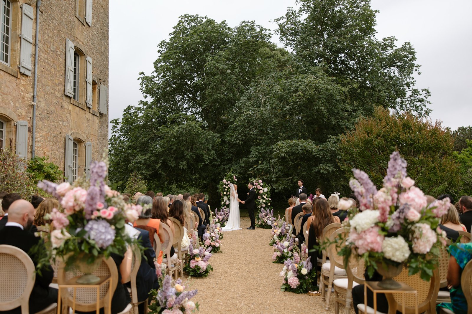 An outdoor wedding ceremony with guests seated on either side of a flower-lined aisle, facing a couple and officiant under a floral arch in front of large trees. Chateau de la Bourlie wedding.