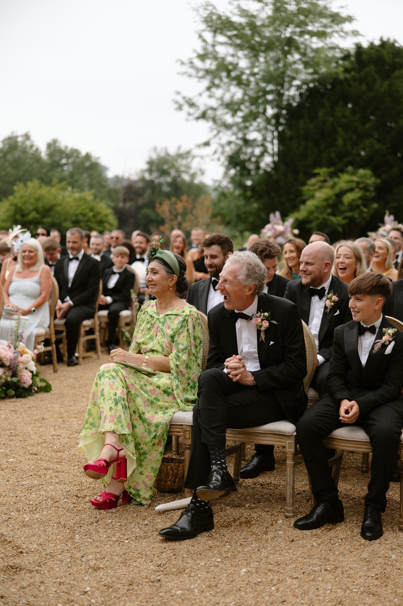 Guests dressed formally are seated outdoors at a wedding ceremony, smiling and facing forward. Trees and greenery are visible in the background. Chateau de la Bourlie wedding.