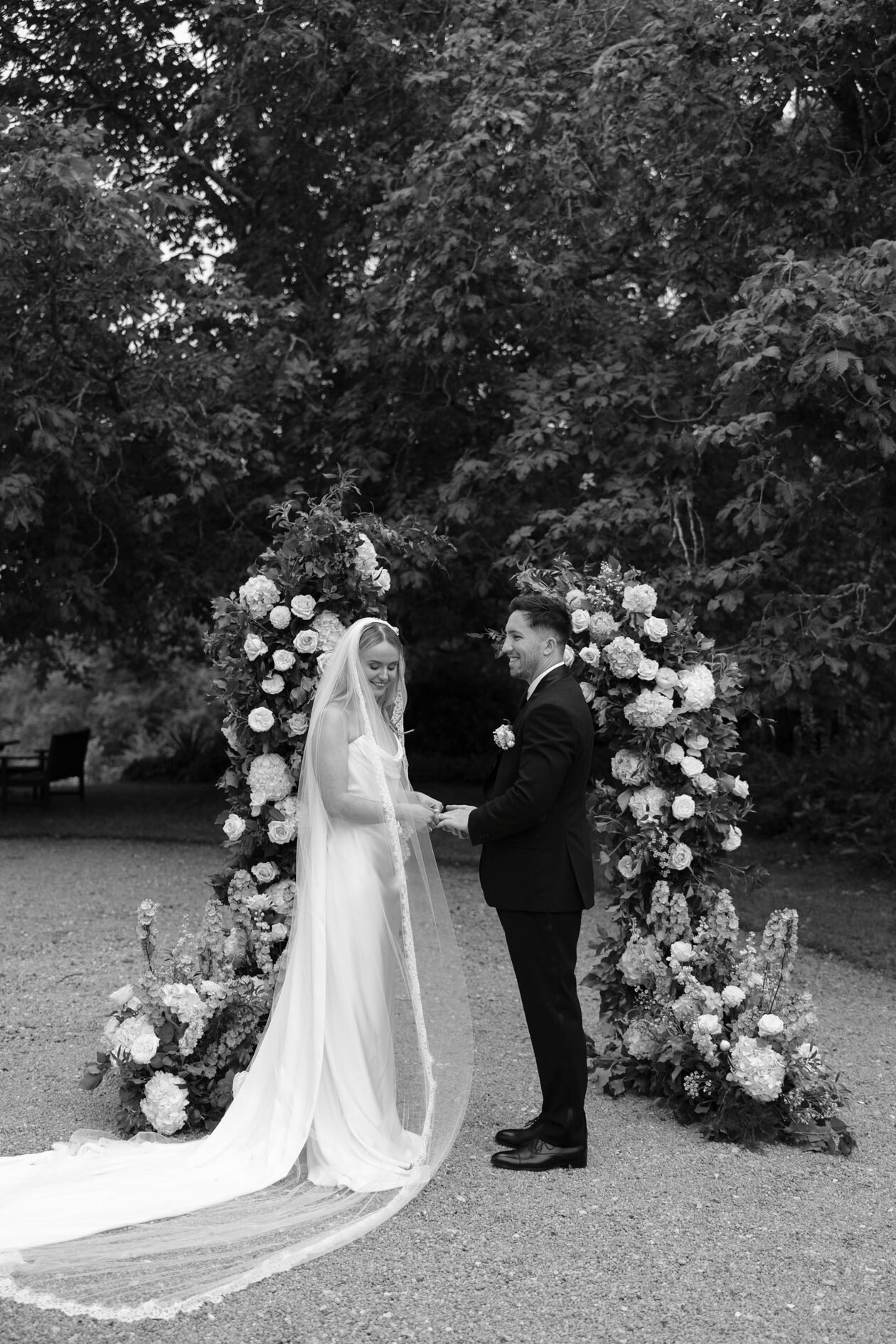 A bride and groom stand holding hands outdoors in front of a floral arch, dressed in wedding attire, surrounded by trees.