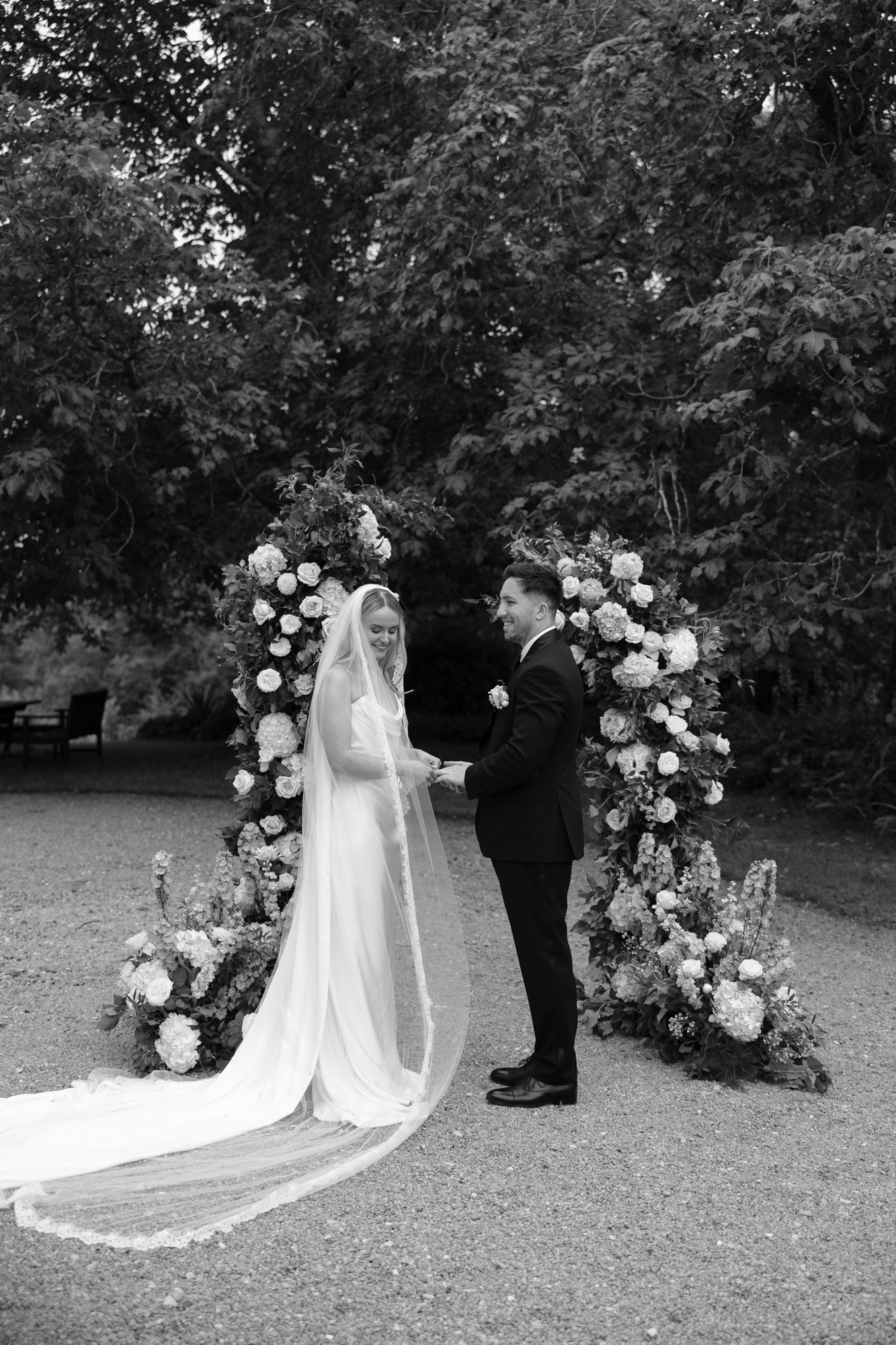 A bride and groom stand holding hands outdoors in front of a floral arch, dressed in wedding attire, surrounded by trees. Chateau de la Bourlie wedding.
