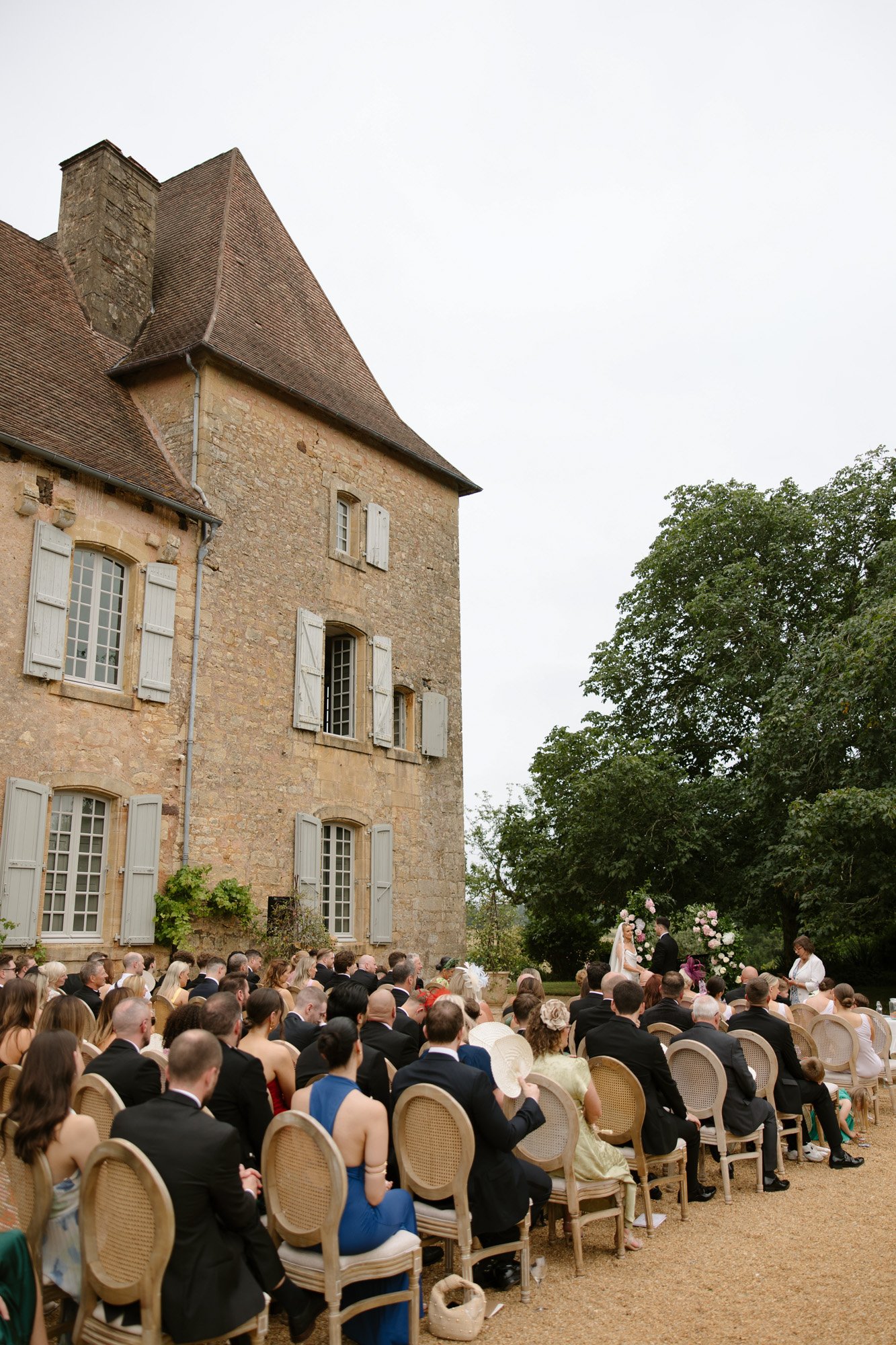 Guests seated outdoors at a wedding ceremony beside a historic stone building with large windows and a sloped roof. Chateau de la Bourlie wedding.