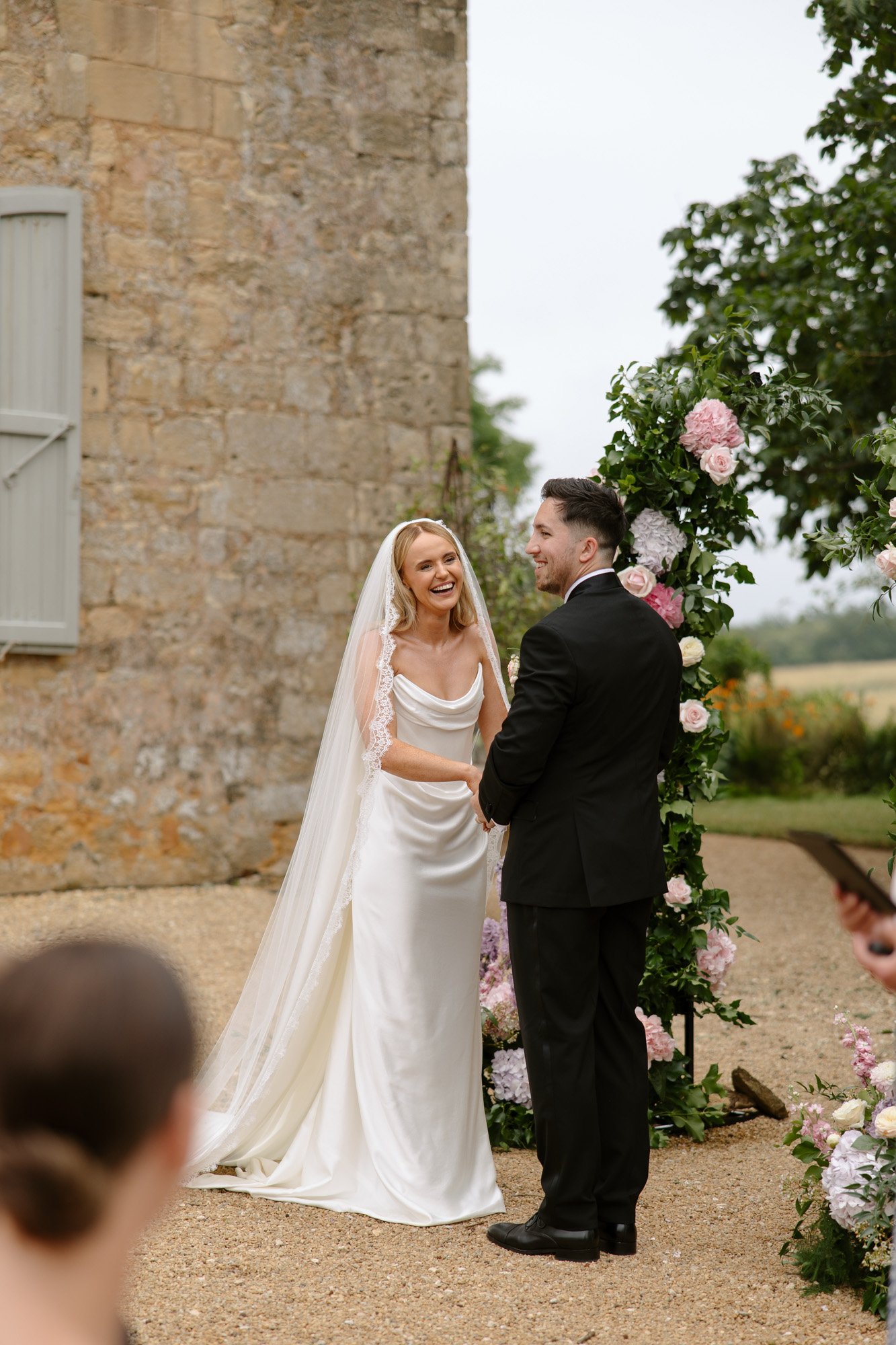 A bride and groom stand facing each other, holding hands and smiling during an outdoor wedding ceremony, with flowers and greenery in the background. Chateau de la Bourlie wedding.
