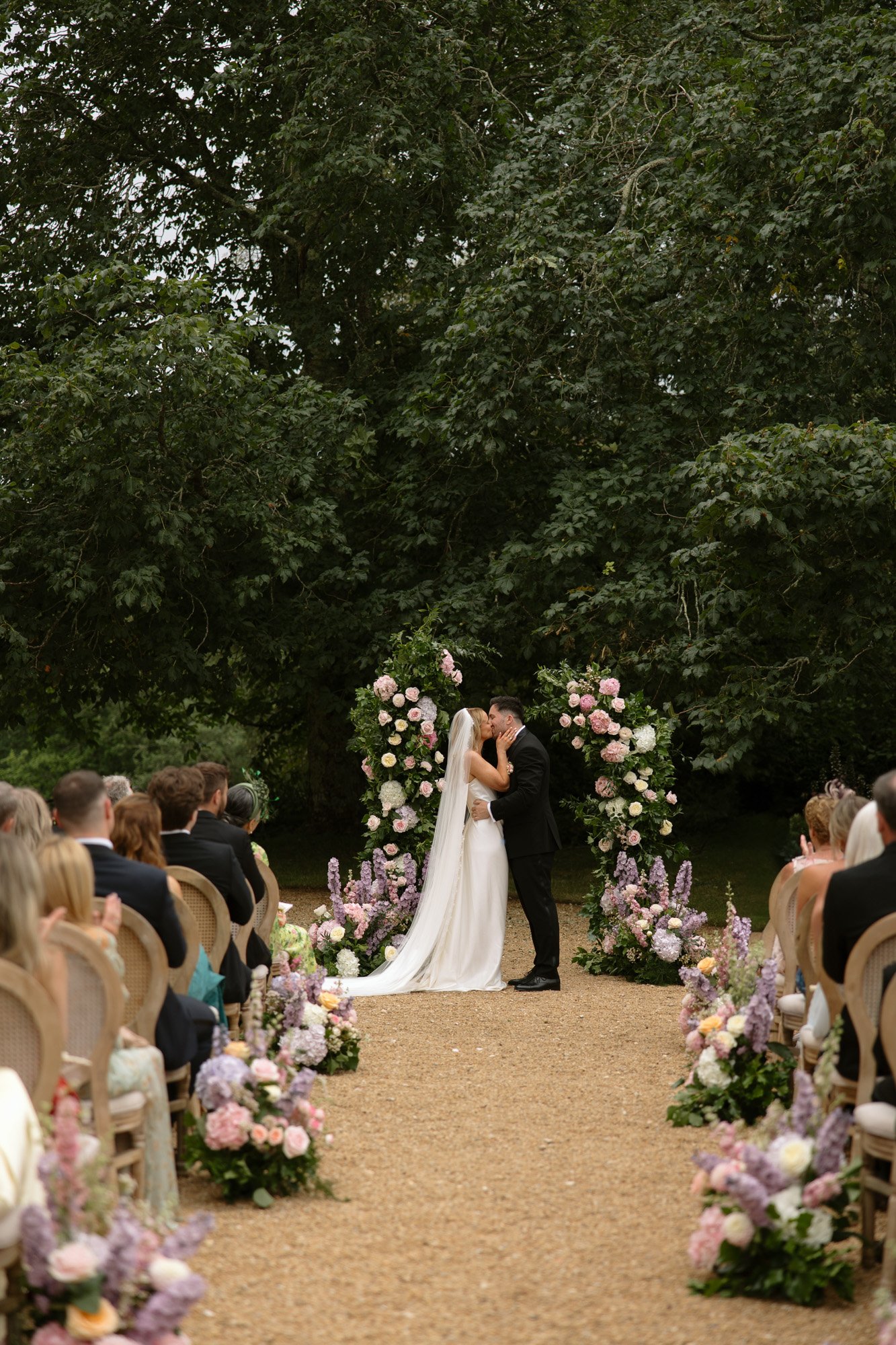 A bride and groom kiss at an outdoor wedding ceremony, standing under a floral arch with guests seated on both sides of the aisle. Chateau de la Bourlie wedding.