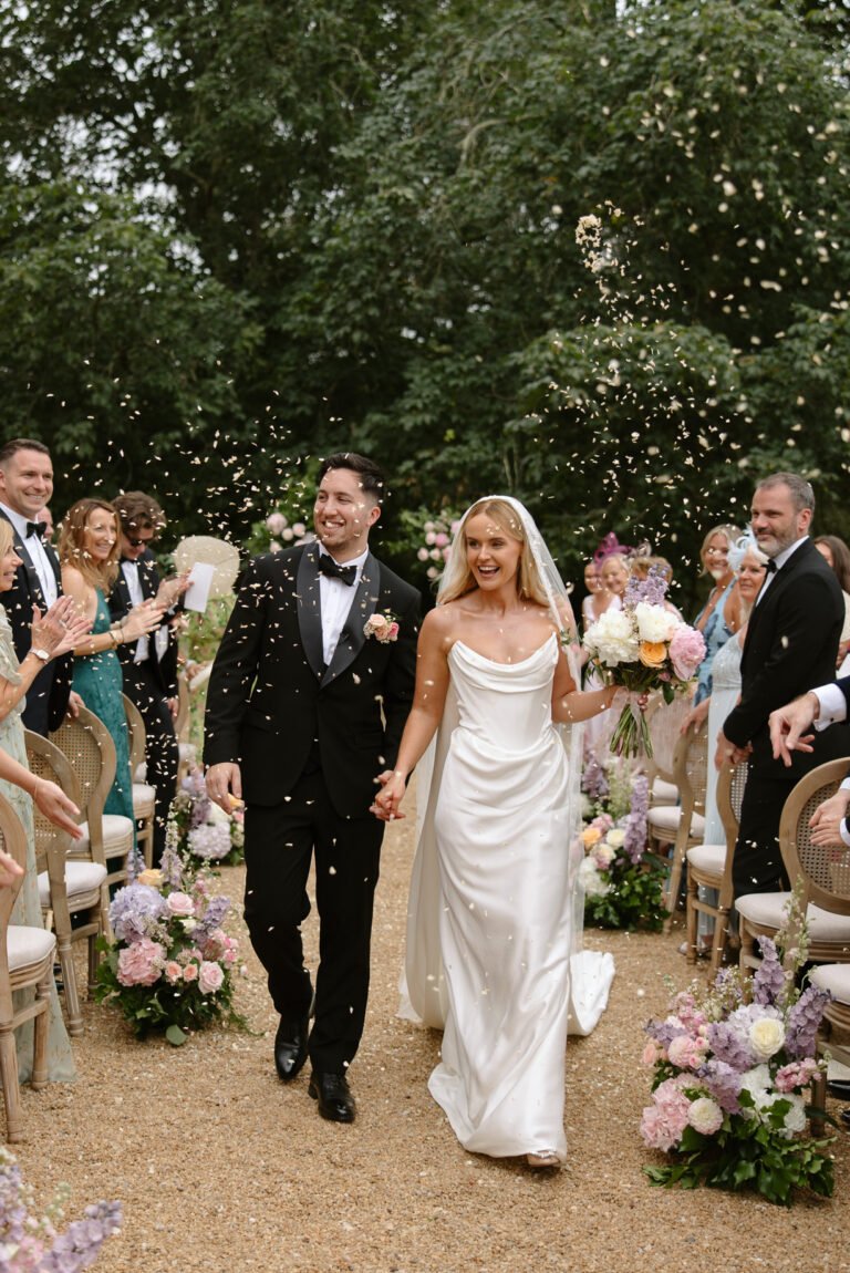 A bride and groom walk down an outdoor aisle, holding hands and smiling, as guests throw confetti. The aisle is decorated with flowers and surrounded by seated guests.