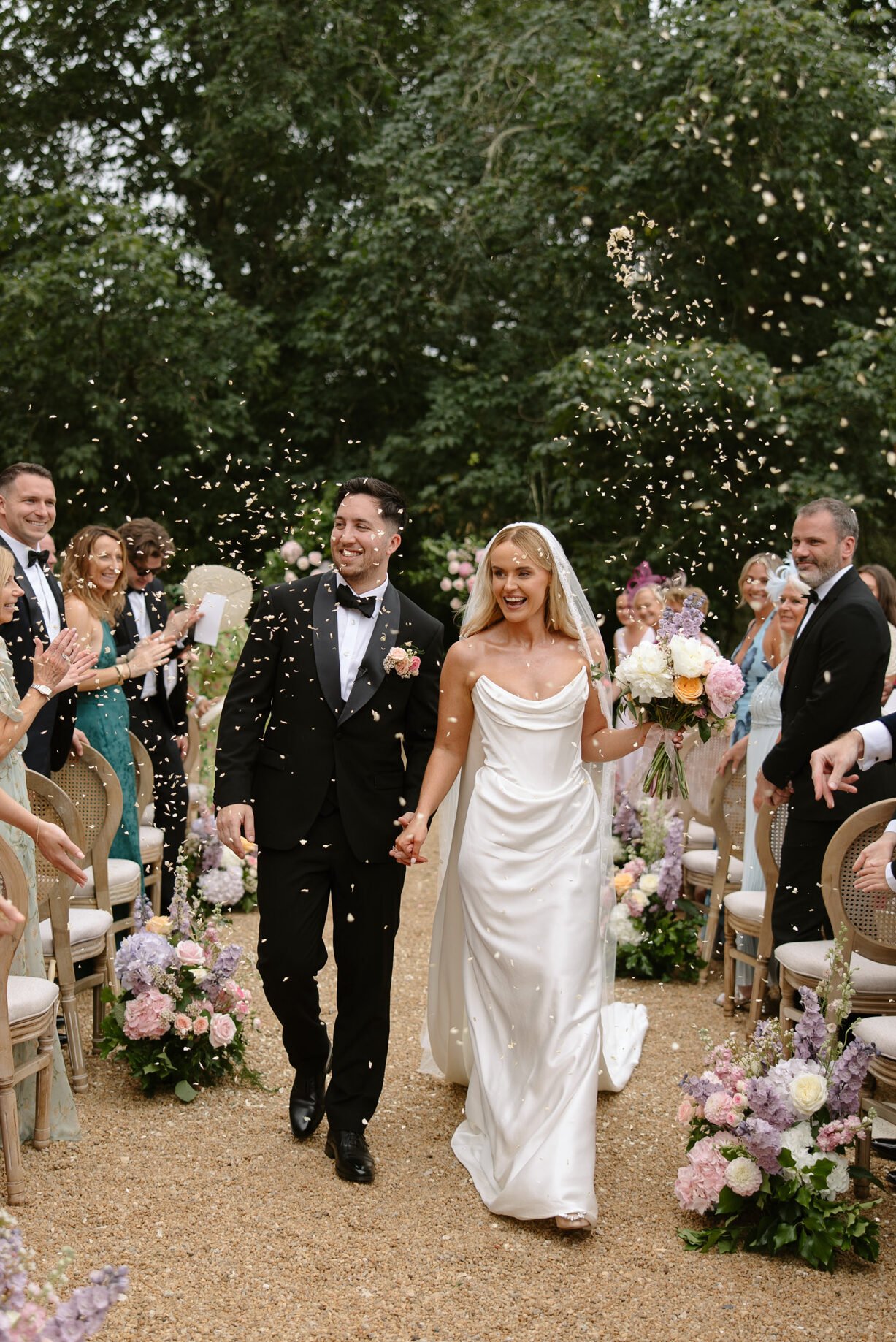 A bride and groom walk down an outdoor aisle, holding hands and smiling, as guests throw confetti. The aisle is decorated with flowers and surrounded by seated guests. France wedding photographer based in Normandy
