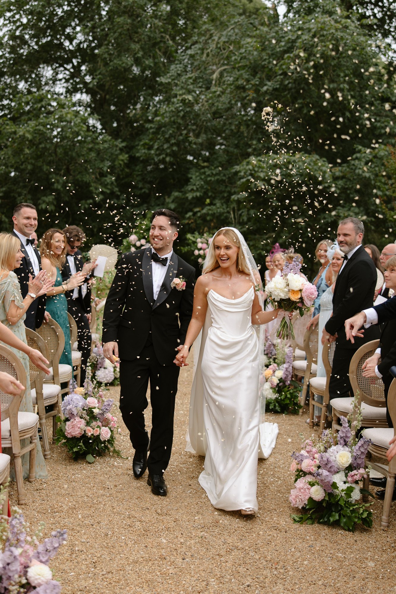 A bride and groom walk down an outdoor aisle, holding hands and smiling, as guests throw confetti. The aisle is decorated with flowers and surrounded by seated guests. Chateau de la Bourlie wedding.