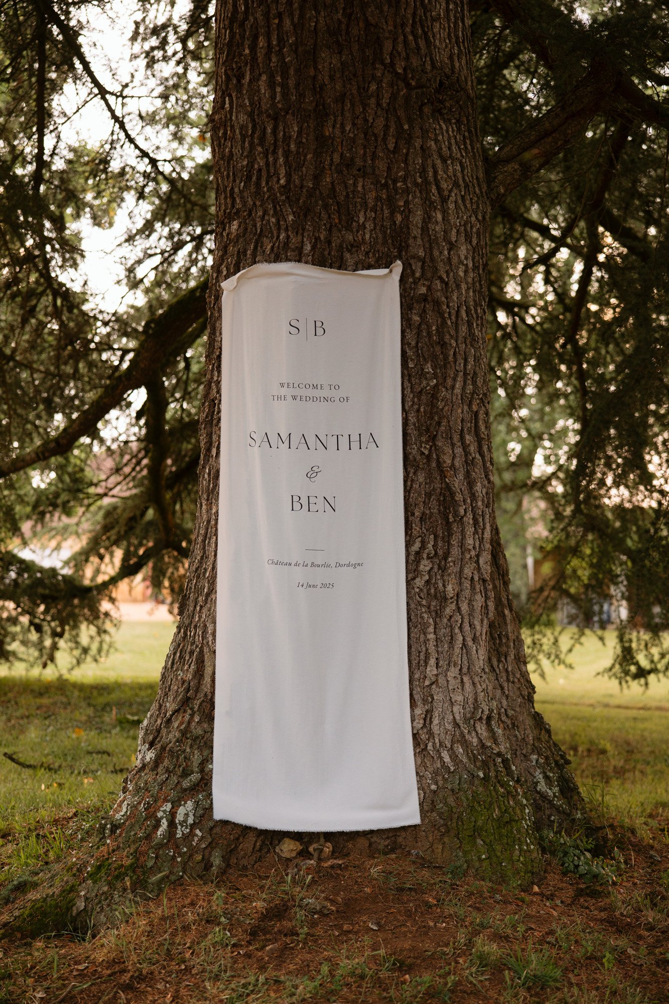 A white wedding banner with black text reading “Welcome to the wedding of Samantha & Ben” is draped on a large tree trunk in an outdoor setting.