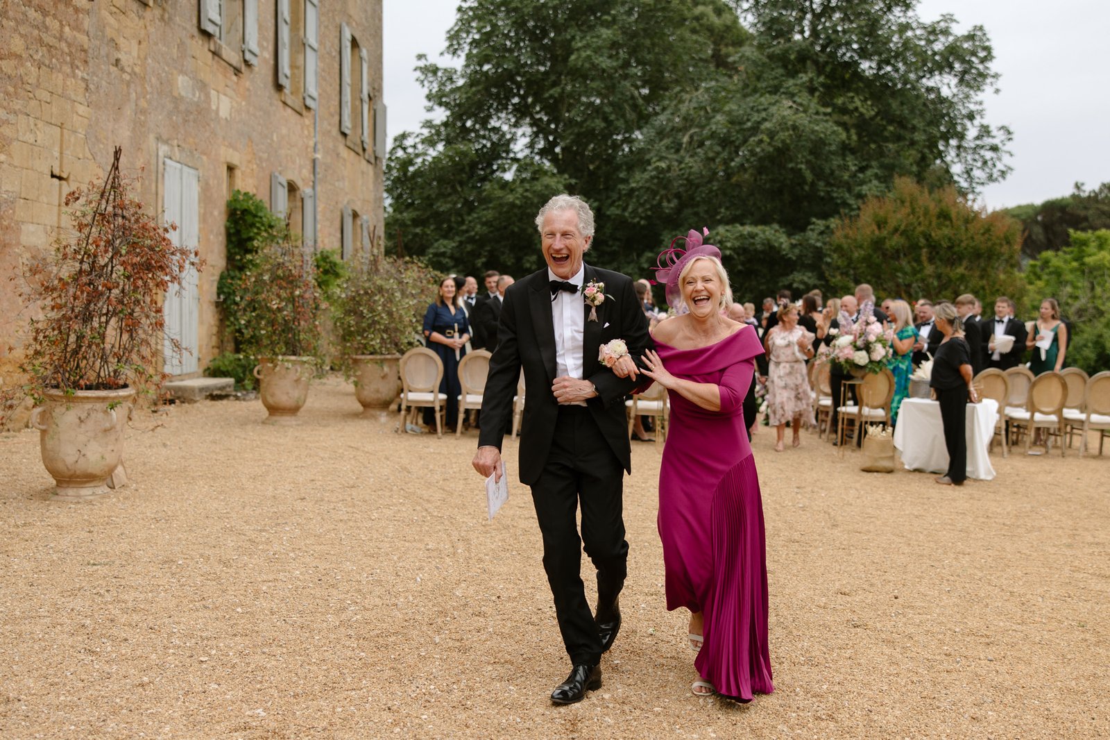 An older man in a tuxedo and an older woman in a bright magenta dress walk arm-in-arm outdoors, smiling, with wedding guests seated behind them. Chateau de la Bourlie wedding.