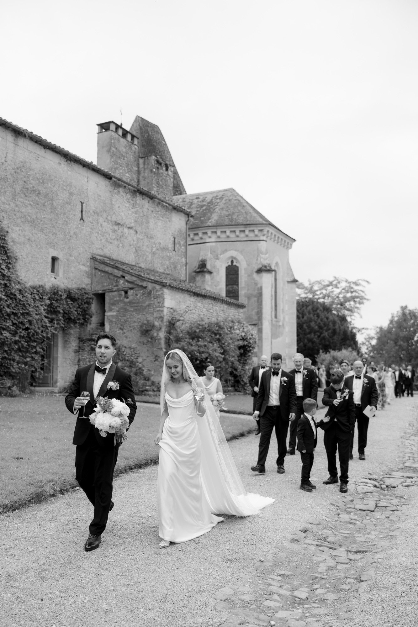 A bride and groom walk outside a stone building, followed by wedding guests dressed in formal attire.