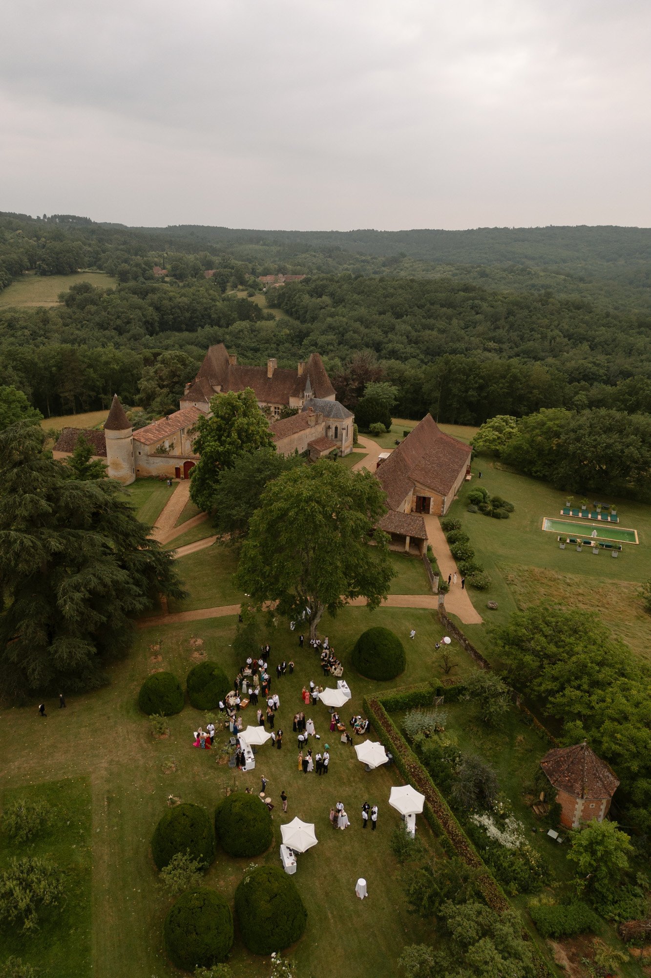 Aerial view of a gathering on the lawn of a large country estate surrounded by trees and gardens, with several buildings and a cloudy sky overhead. Chateau de la Bourlie wedding.
