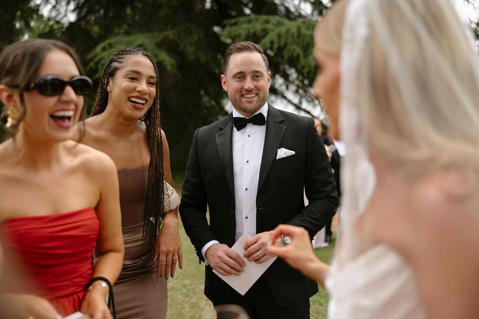 Three guests and a bride converse outdoors; two women smile, one in red and one in taupe, a man in a tuxedo holds an envelope, and trees are visible in the background.