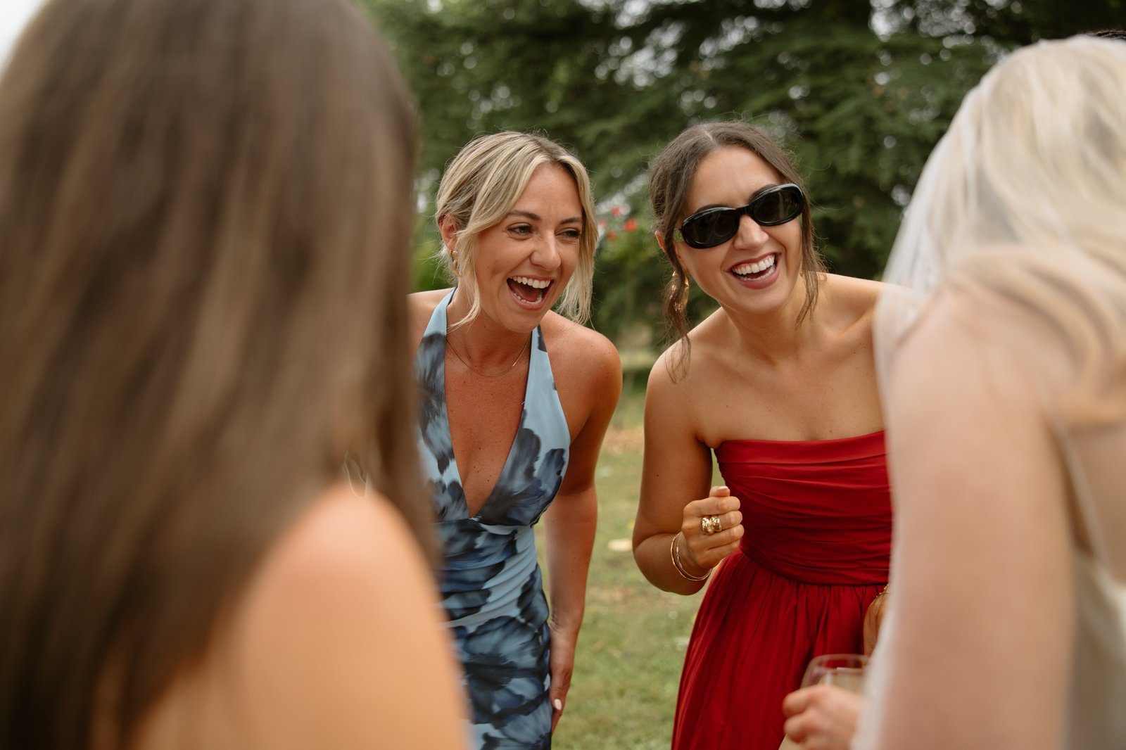 Two women, one in a blue dress and one in a red dress with sunglasses, laugh together outdoors with two other people partially visible in the foreground.