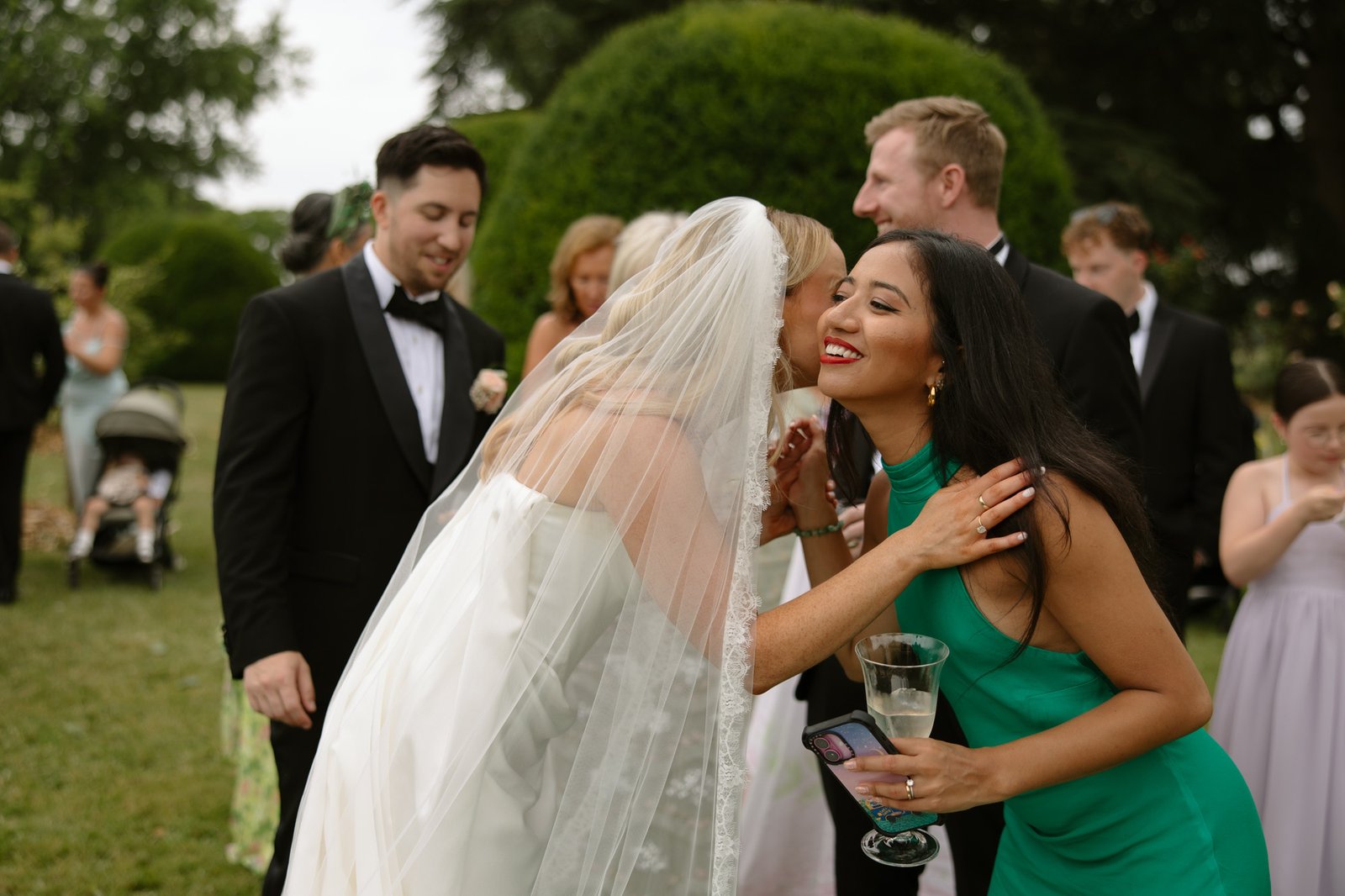A bride in a veil kisses a woman in a green dress on the cheek at an outdoor event, with other guests mingling in the background.