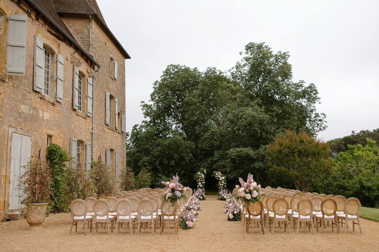 Outdoor wedding ceremony setup with rows of chairs facing a floral arrangement, next to a stone building and greenery in the background. Chateau de la Bourlie wedding.