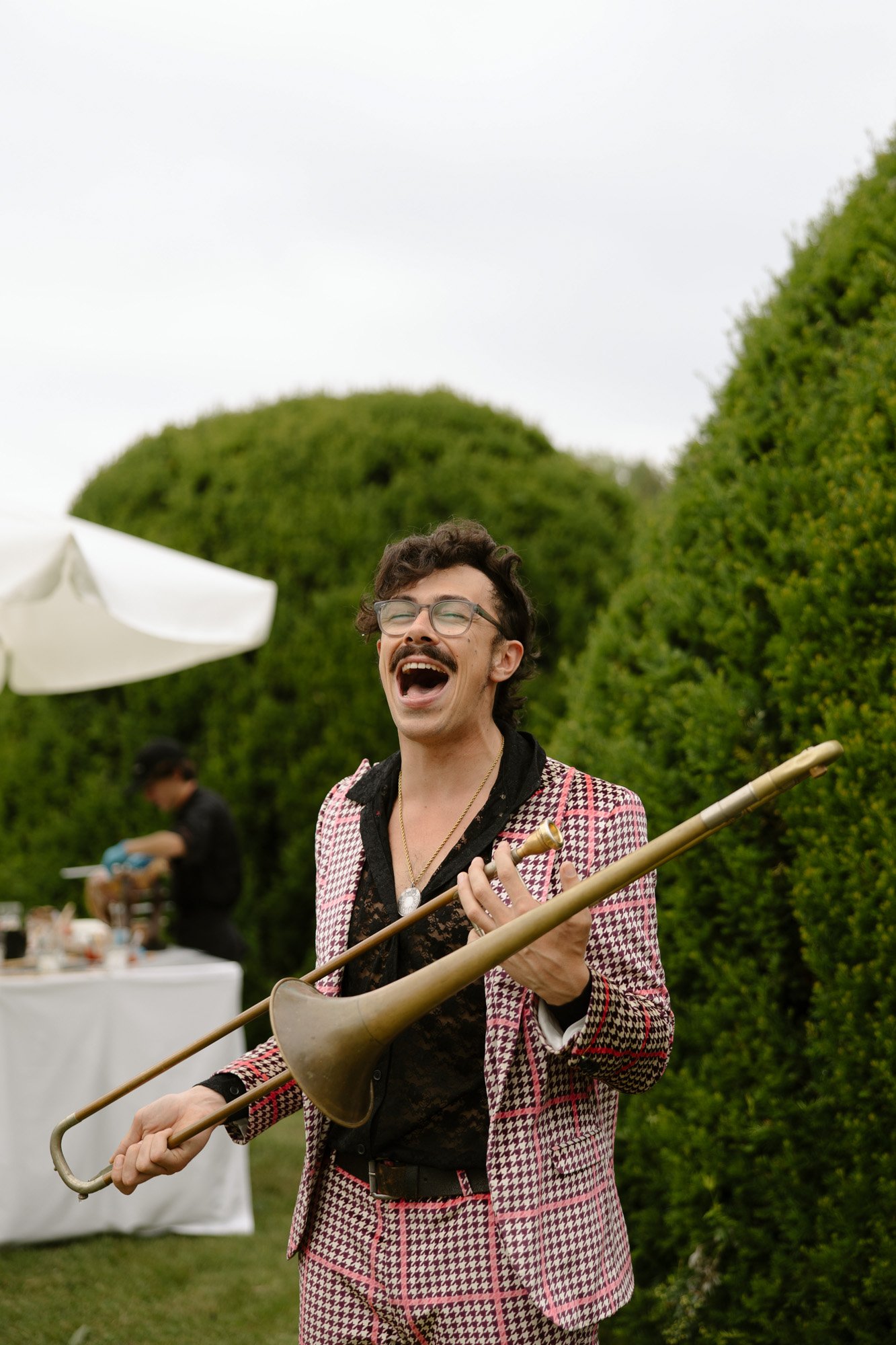 A person wearing a pink plaid suit and glasses stands outdoors, holding a trombone and smiling with mouth open. There are green bushes and a table with food in the background.