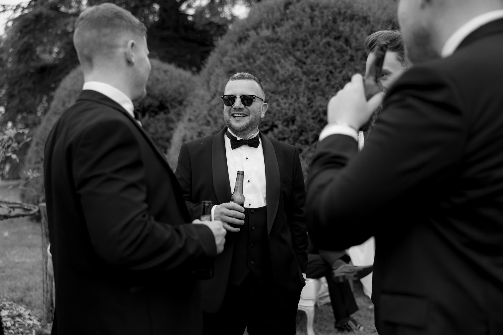 Four men in suits, one holding a bottle and wearing sunglasses, stand talking and smiling outdoors in a garden setting.