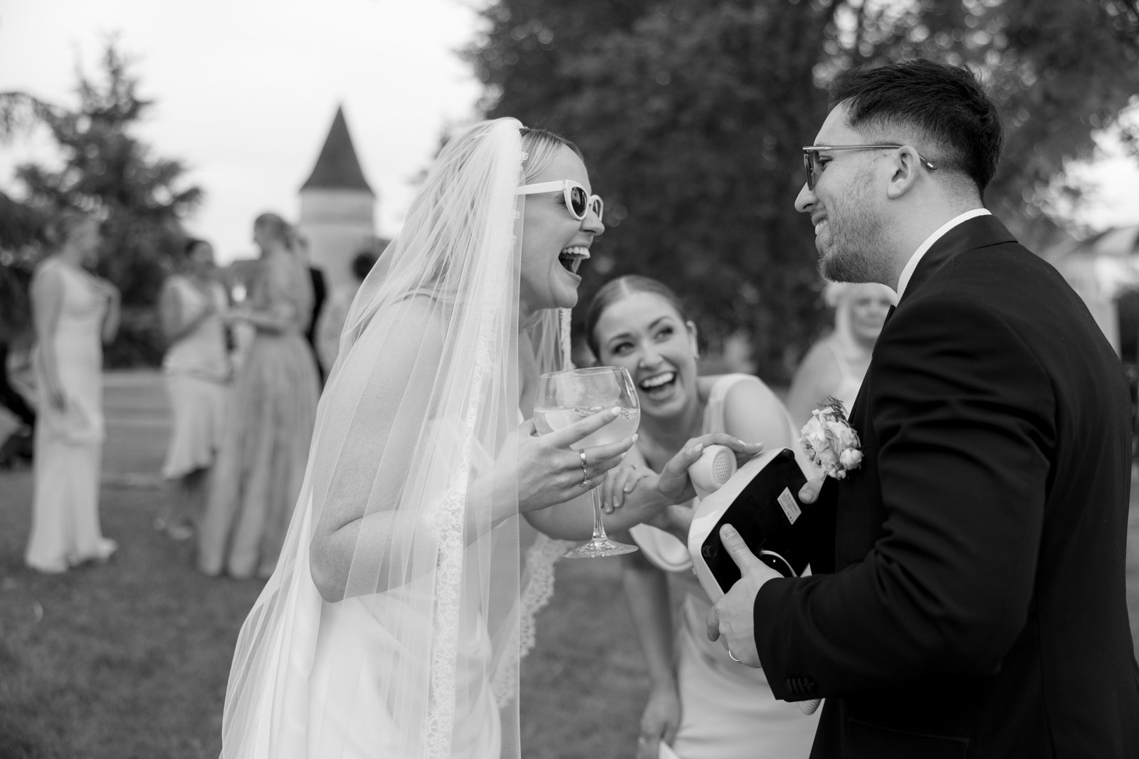 A bride in a veil and sunglasses laughs with a man in a suit while holding a drink at an outdoor gathering, with guests in the background.