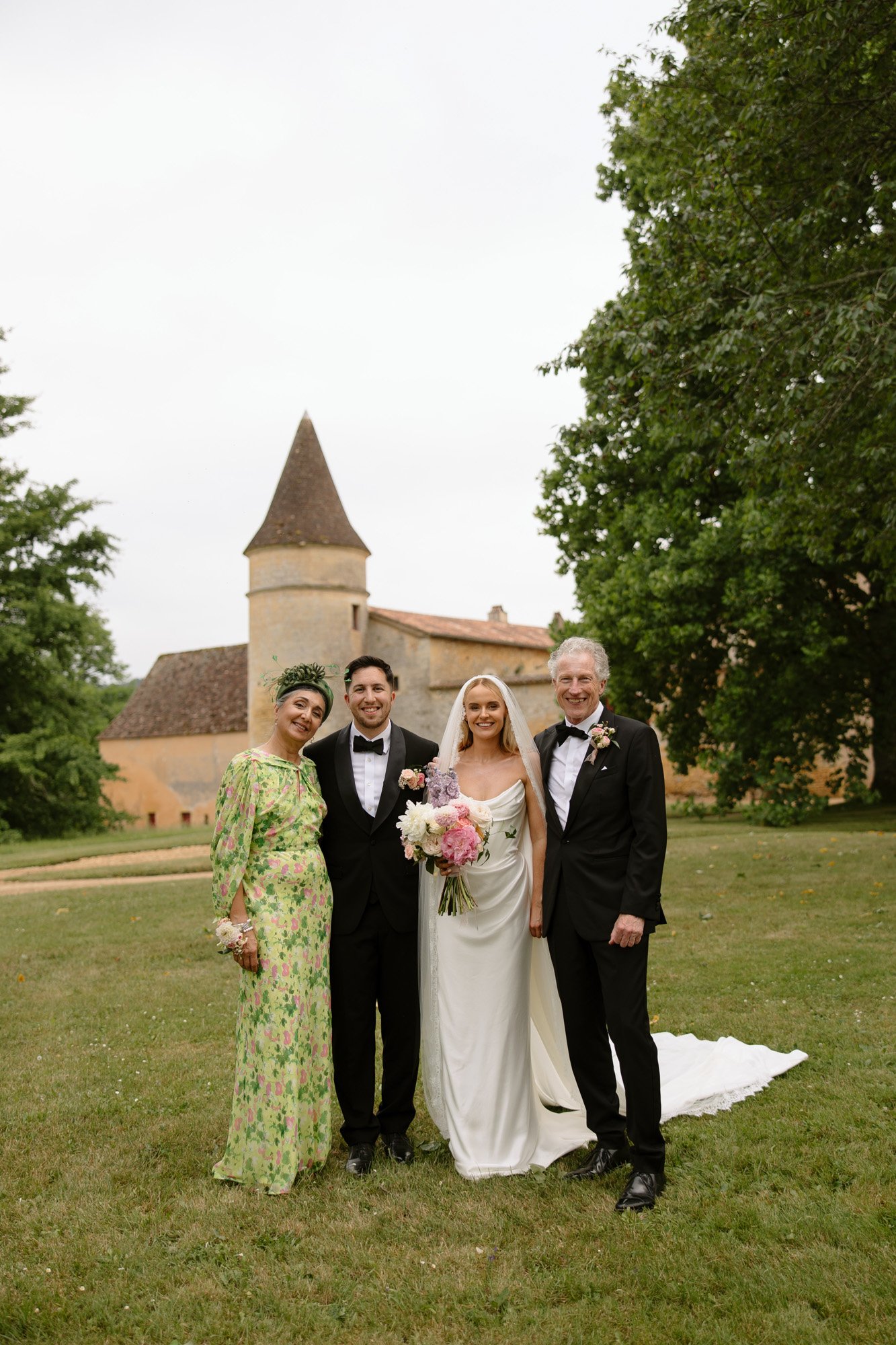 Four people, including a bride and groom in wedding attire, stand together outdoors in front of a historic stone building and trees, posing for a formal photo.